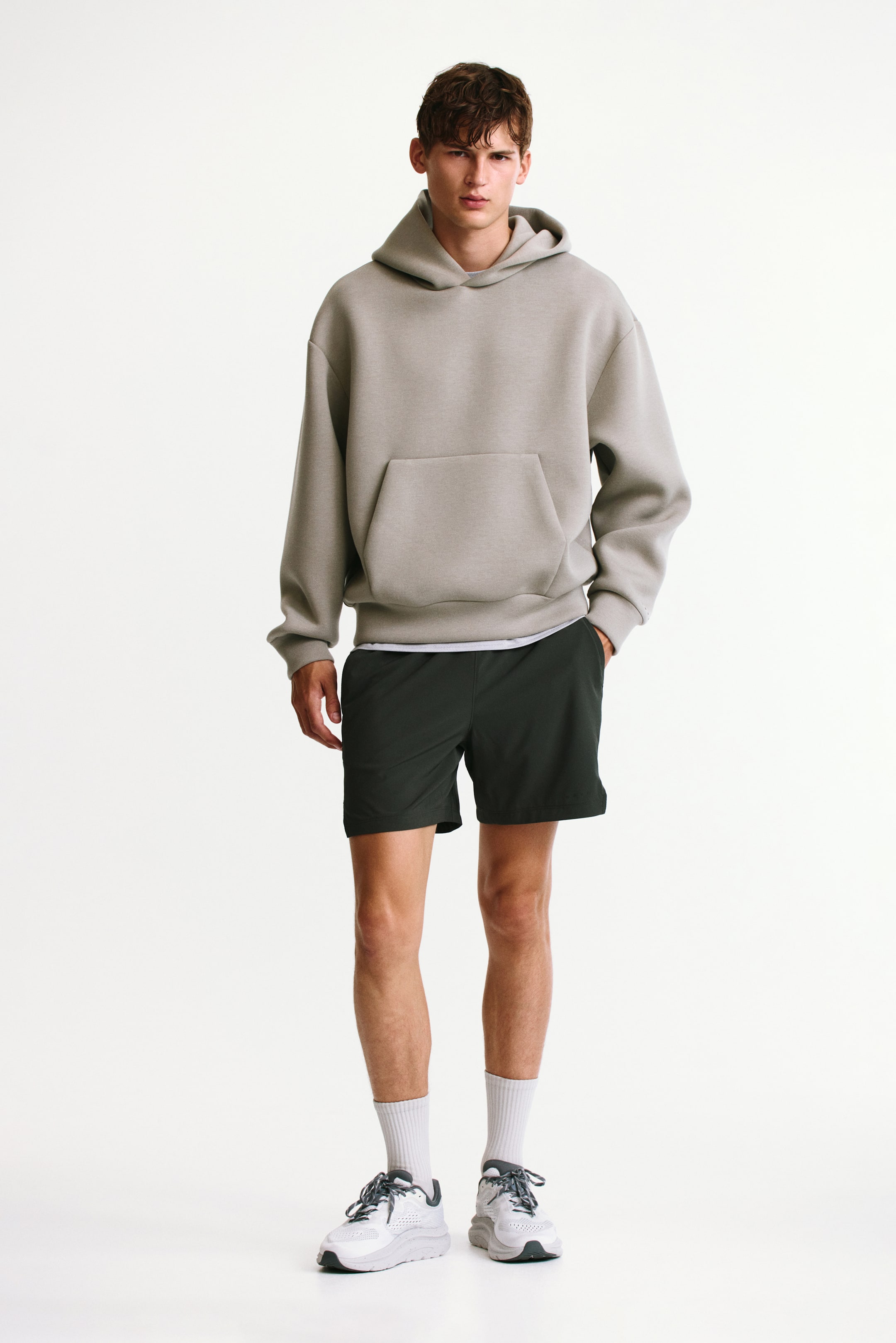 Xem ảnh lớn hơn: Một chiếc hoodie dáng rộng màu xám nhạt, nổi bật với túi kangaroo phía trước và vai trễ, đang được người mẫu mặc, phối cùng quần short thể thao màu xanh lá đậm. Người mẫu đứng với một tay đặt trong túi quần short, kết hợp cùng tất cổ ngắn màu trắng và giày sneaker thể thao màu xám.