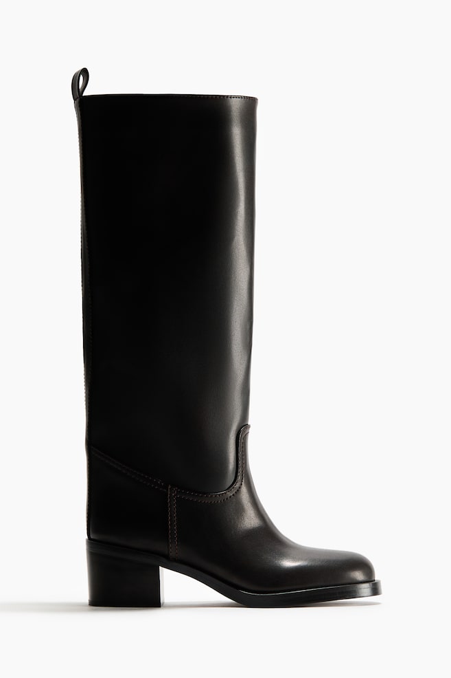A black boot is presented in a side profile, featuring rounded toes, a block heel, and a knee-high shaft with a loop at the back. - 1
