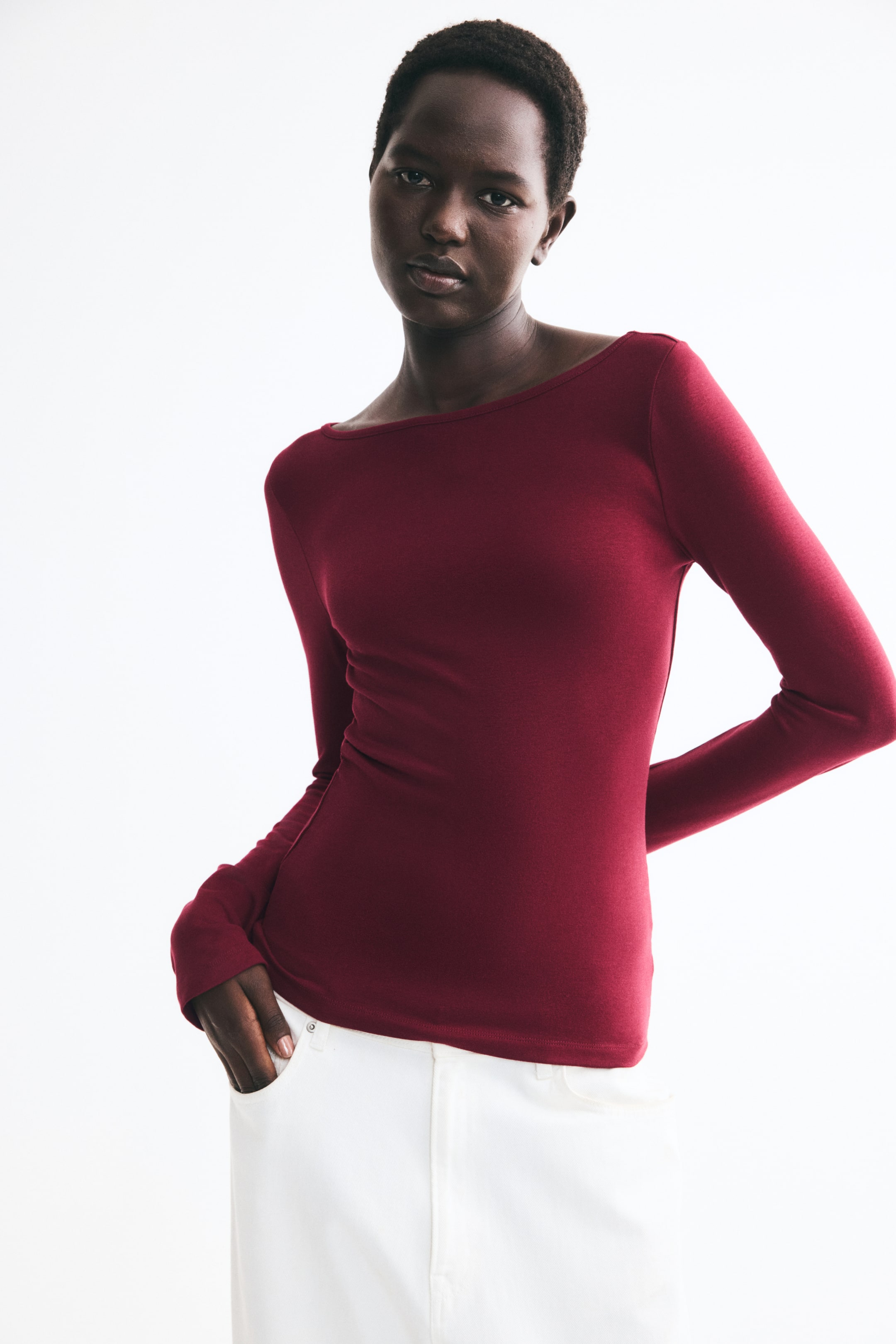 View larger image: A dark red, long-sleeved top in a fine knit is being worn by the model, featuring a wide boat neckline and a straight hem. The top is styled with white trousers, with the model placing one hand inside a front pocket.