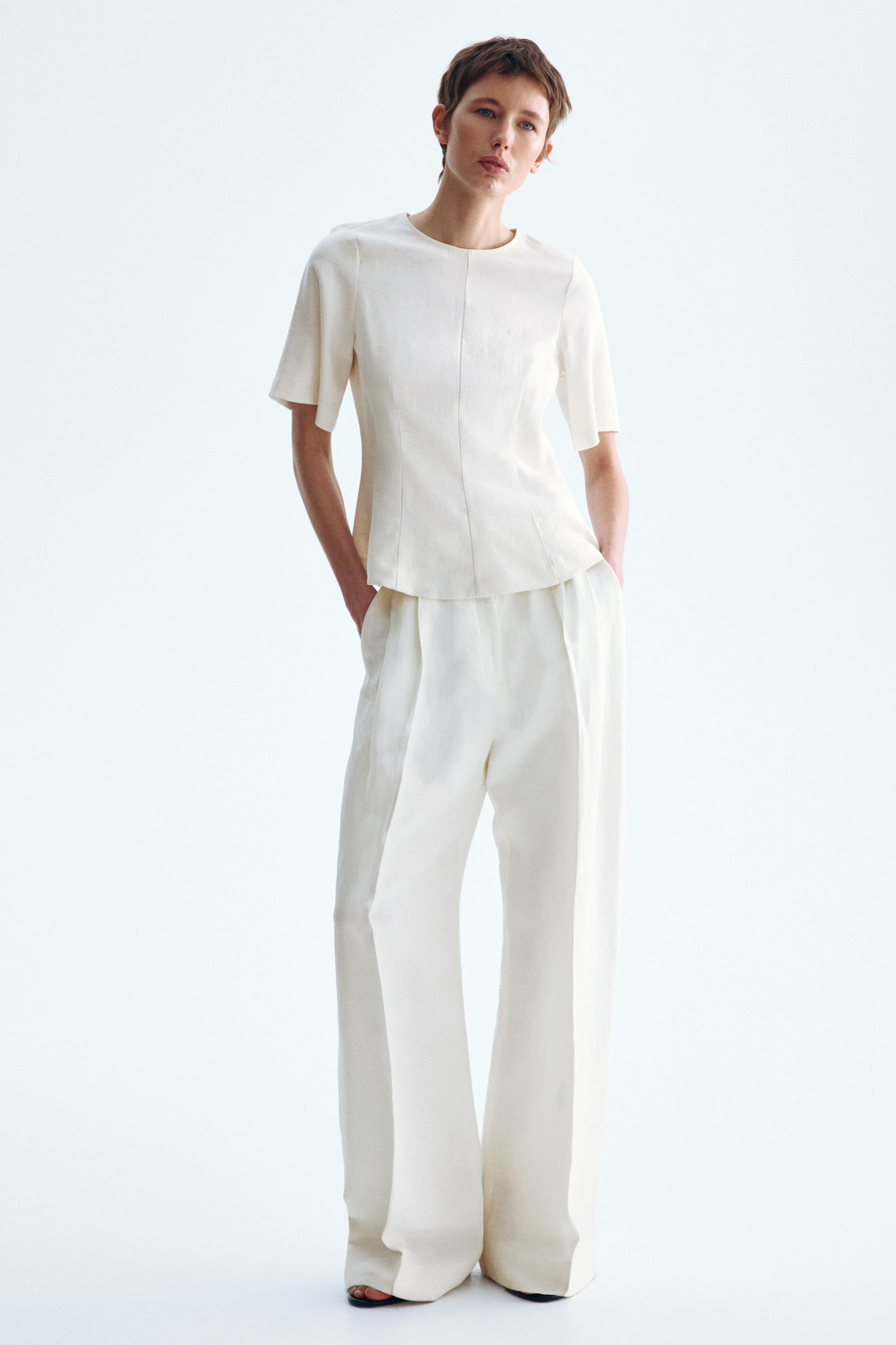 View larger image: A cream-colored short-sleeved top and matching wide-leg pants are displayed from a front-facing angle. The top features a round neckline and vertical seams, while the high-waisted pants showcase front pleats.