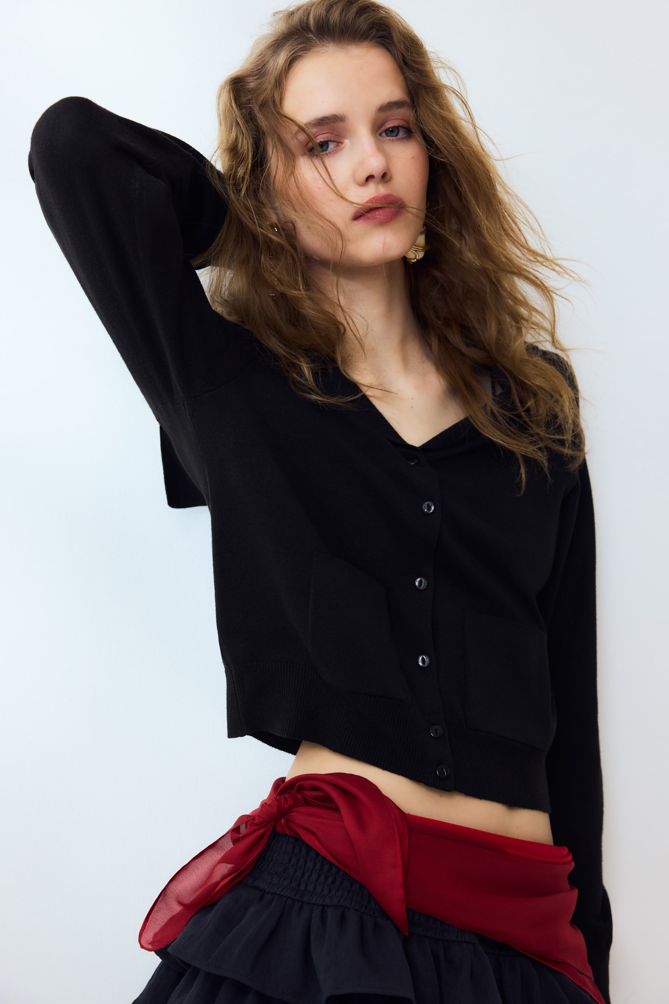 הצגת תמונה גדולה יותר: קרדיגן שחור עשוי סריג דק נלבש על ידי הדוגמנית, עם מחשוף V, צווארון מלחים, כפתורים בחזית וכיסים קדמיים עם טלאים. הדוגמנית מצולמת עם יד אחת מורמת, חושפת את השרוולים המוגמרים בצלעות ואת השוליים הקצרים של הקרדיגן.