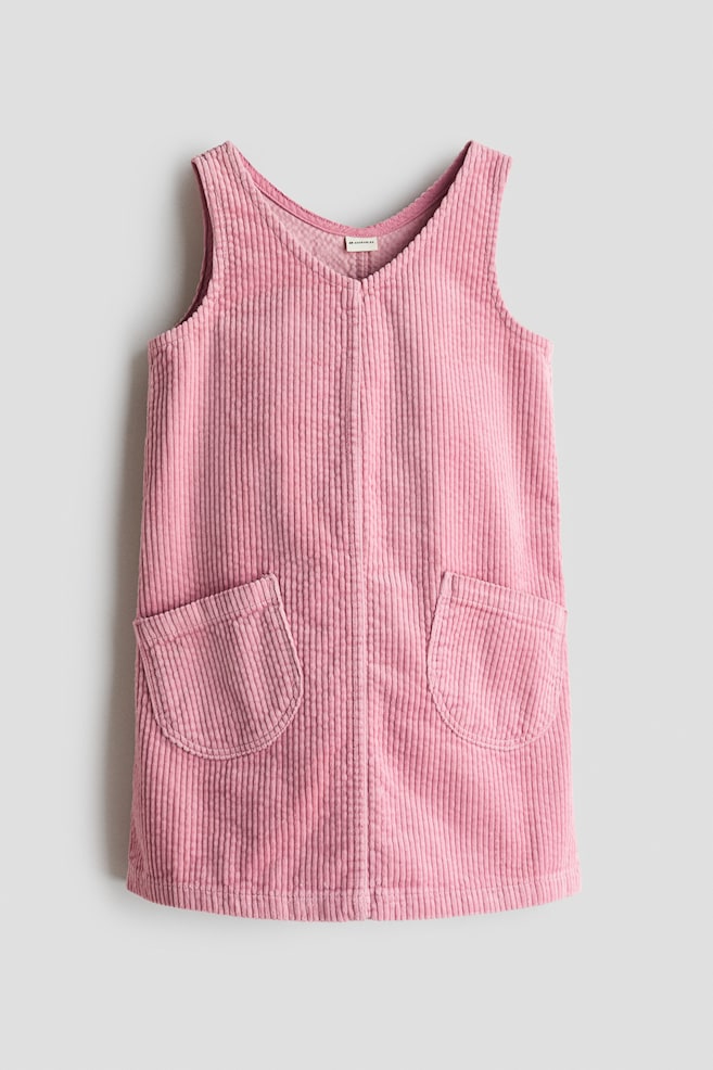 A pink corduroy pinafore dress is shown from the front, featuring a V-shaped neckline and a loose fit. It is displaying two rounded patch pockets on the front and its characteristic vertical corduroy ridges. - 1