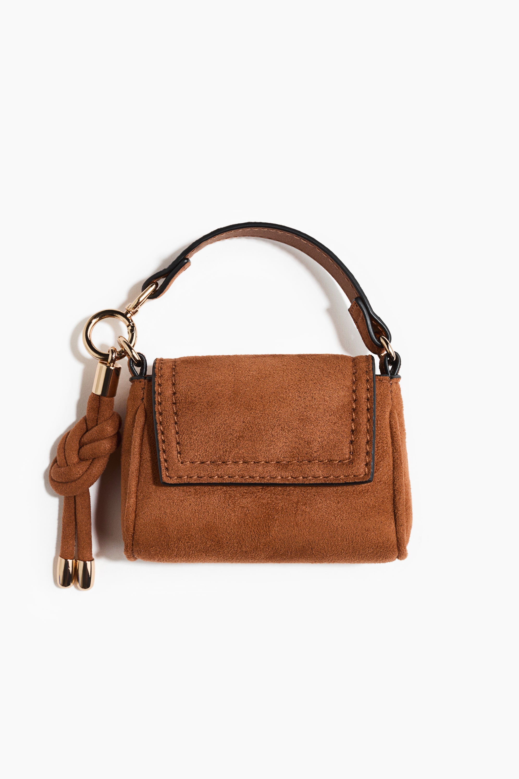 Ver imagen más grande: Un mini bolso marrón de efecto ante se presenta de frente, con cierre de solapa y costuras visibles en la parte frontal. Lleva un asa superior negra sujeta con herrajes en tono dorado, y un adorno anudado a juego con extremos rematados en dorado que cuelga por la izquierda.