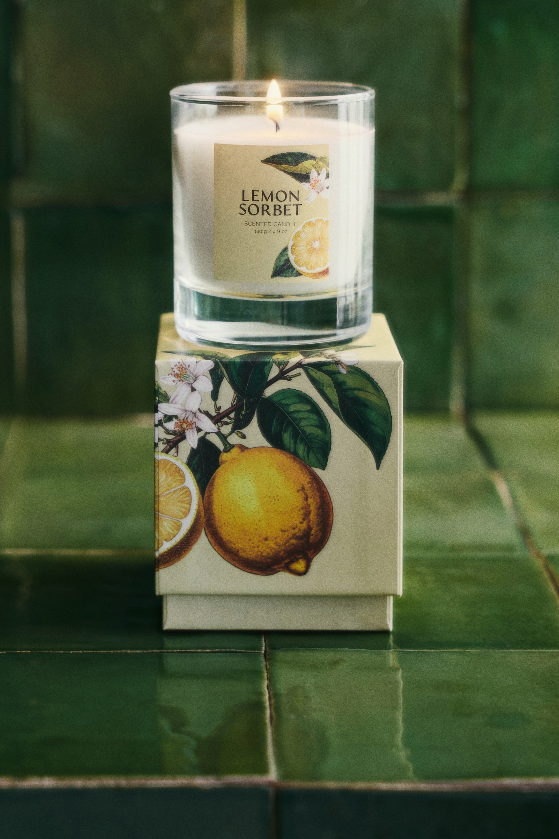 Zobraziť väčší obrázok: Sviečka s vôňou „Citrónový sorbet“, umiestnená v priehľadnej sklenenej nádobe, sedí na svojom zodpovedajúcom svetložltom darčekovom balení. Etiketa sviečky aj krabička obsahujú botanické ilustrácie citrónov, citrónových plátkov a bielych kvetov, zatiaľ čo knôt sviečky horí malým plamienkom.