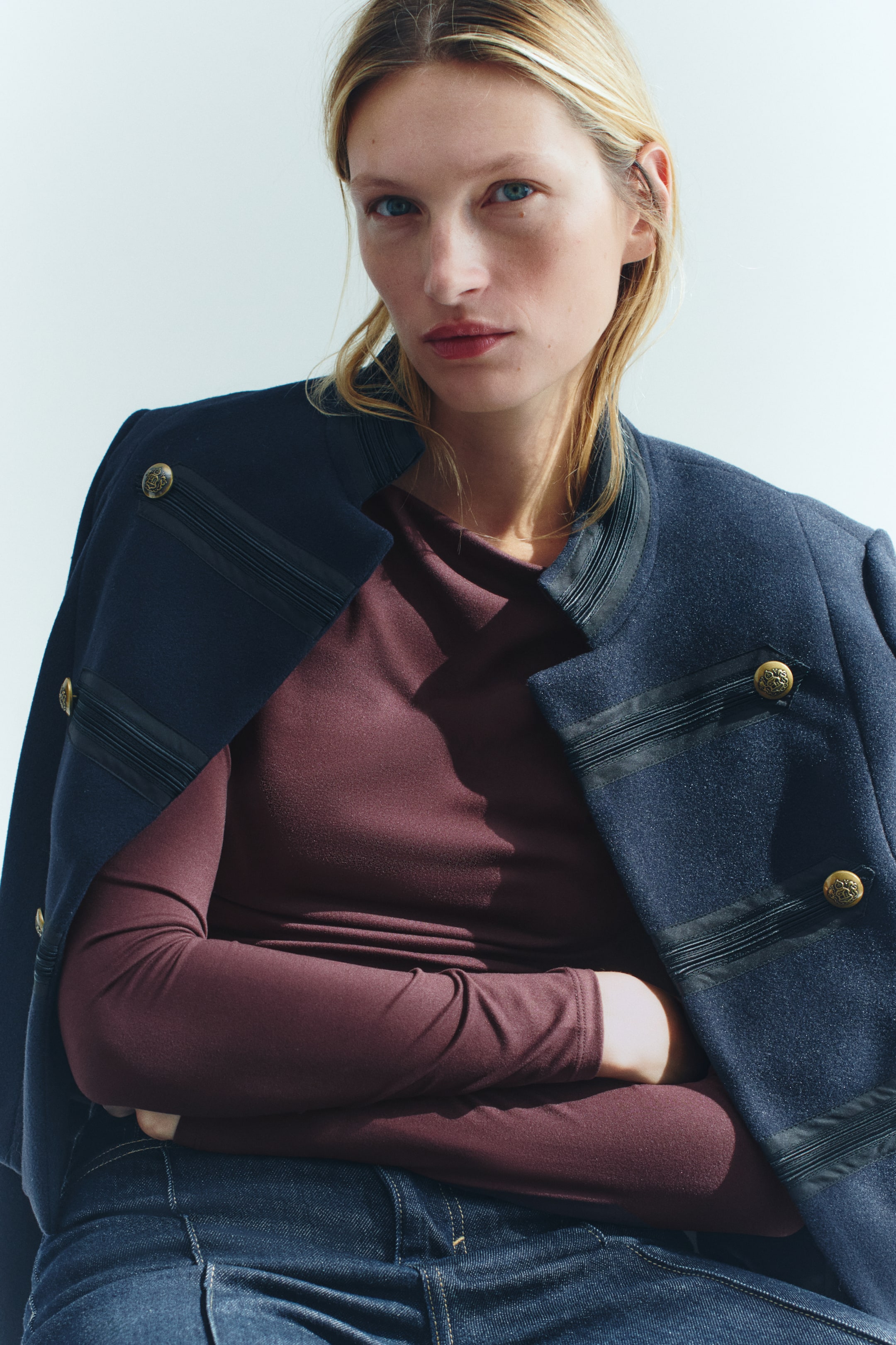 View larger image: A fitted top in dark plum stretch jersey featuring a boat neck and long sleeves is being worn by the model with arms crossed. A navy blue jacket with gold buttons is draped over the shoulders, layering over the top.