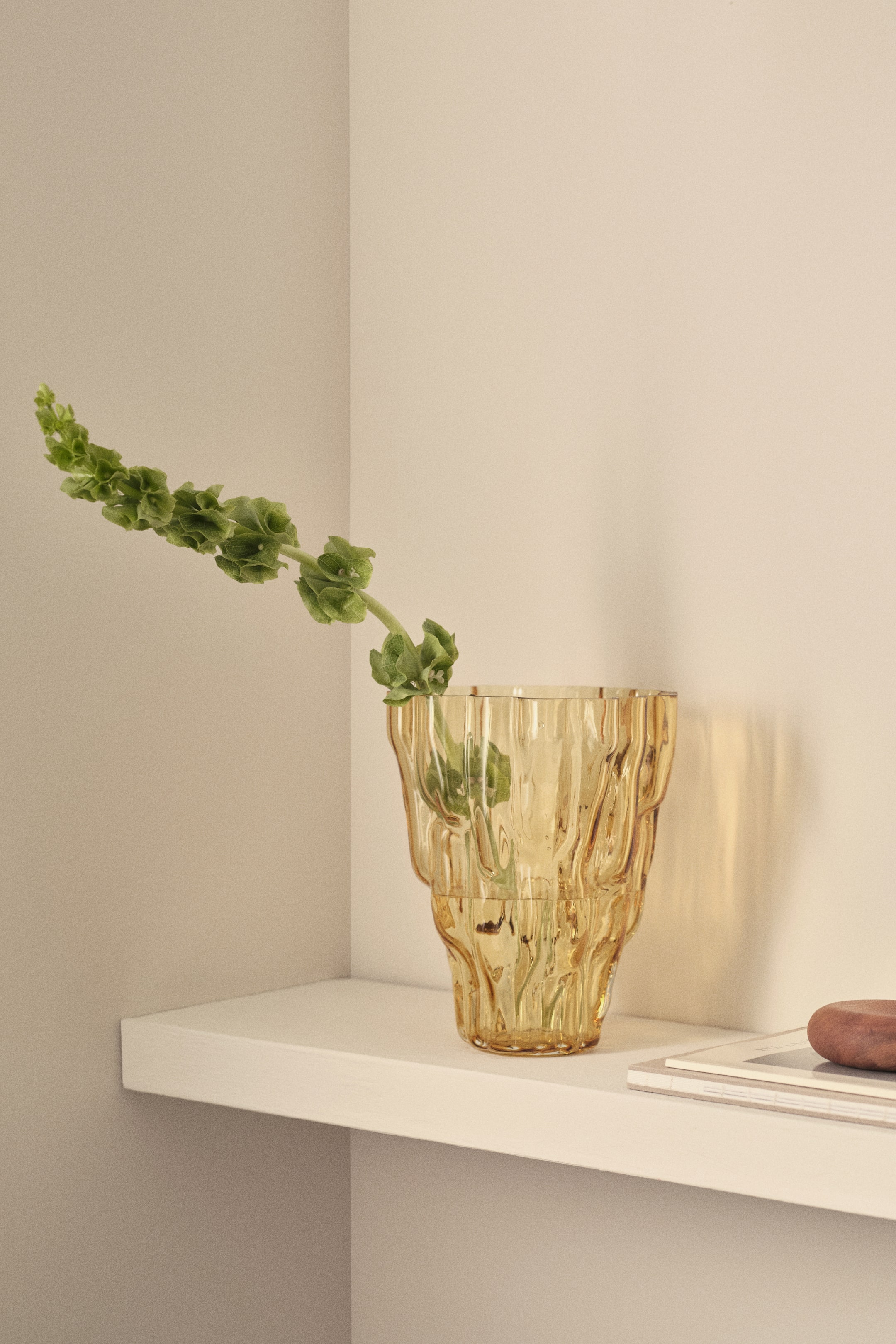 View larger image: An amber glass vase features an asymmetric, sculptural design with irregular, wavy textures. A green plant stem is emerging from its opening.