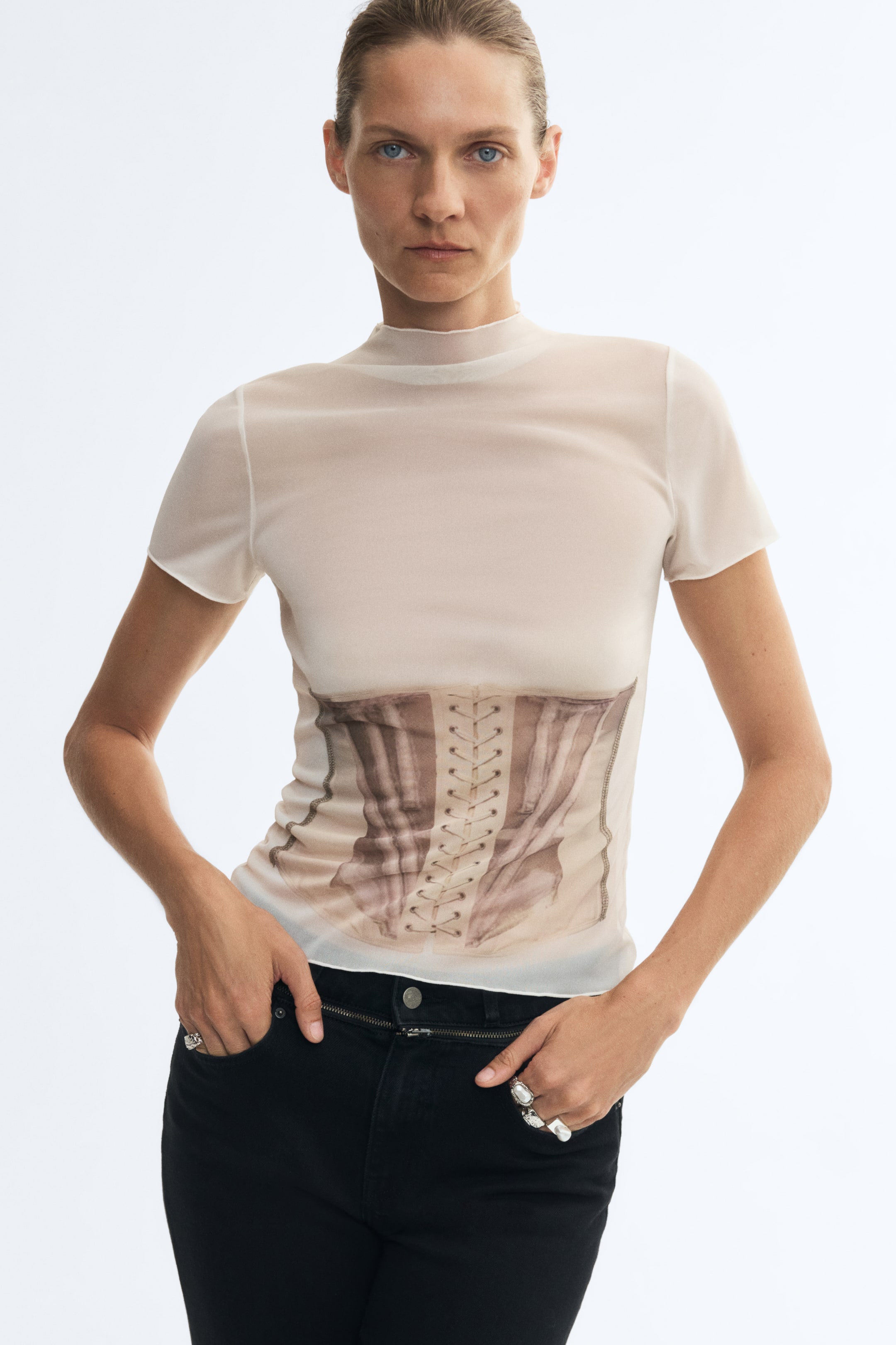 Visa större bild: En krämfärgad mesh-topp med ståkrage, korta ärmar och ett tryckt korsettmotiv framtill bärs av modellen. Modellen stylar toppen med svarta jeans och vilar händerna i framfickorna.