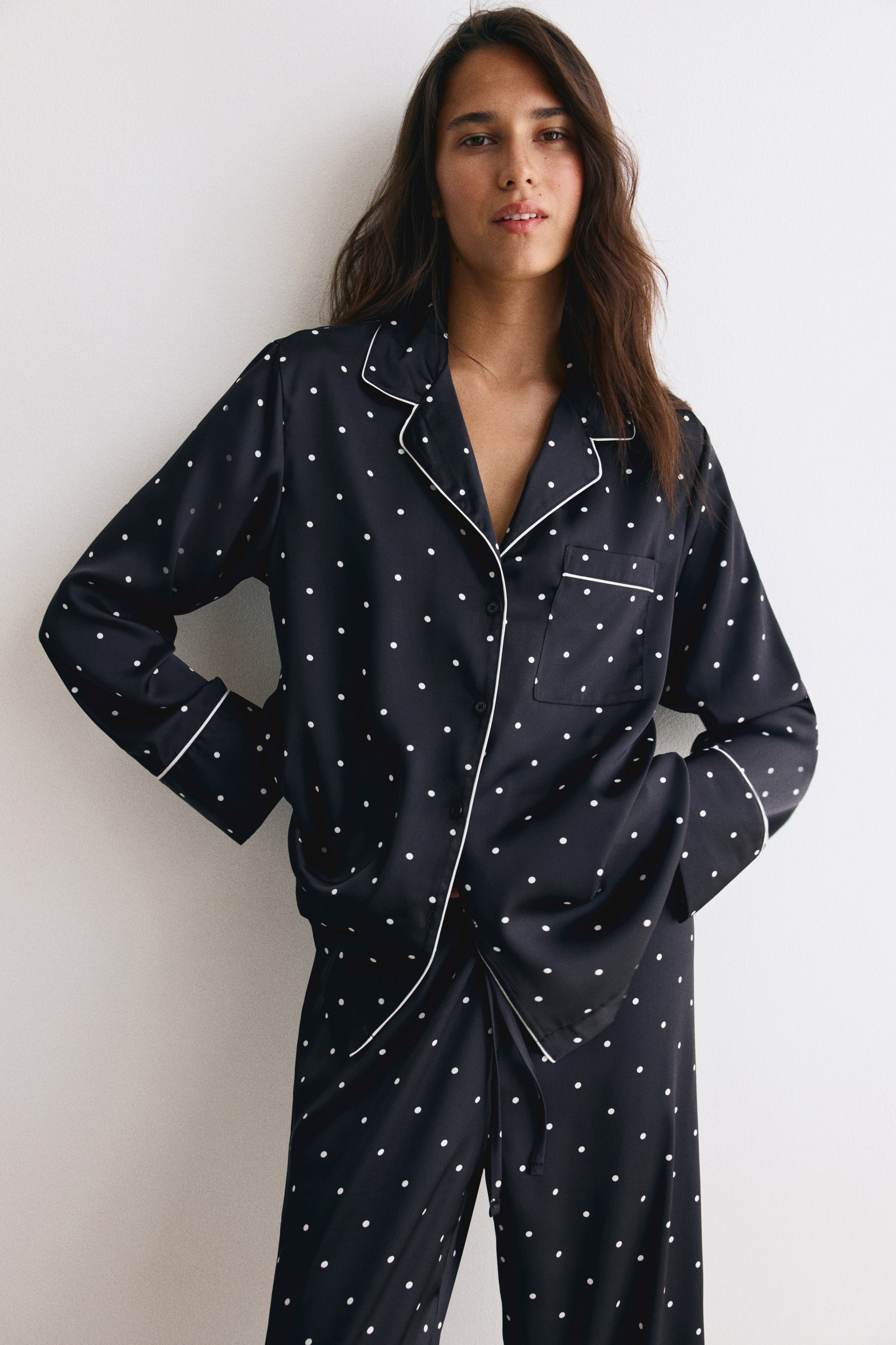 View larger image: A satin pyjama set in black with white polka dots and contrast white piping is being worn by the model. The long-sleeved shirt features a resort collar, button-down front, and chest pocket, while the matching wide-leg pants feature a visible drawstring waist.