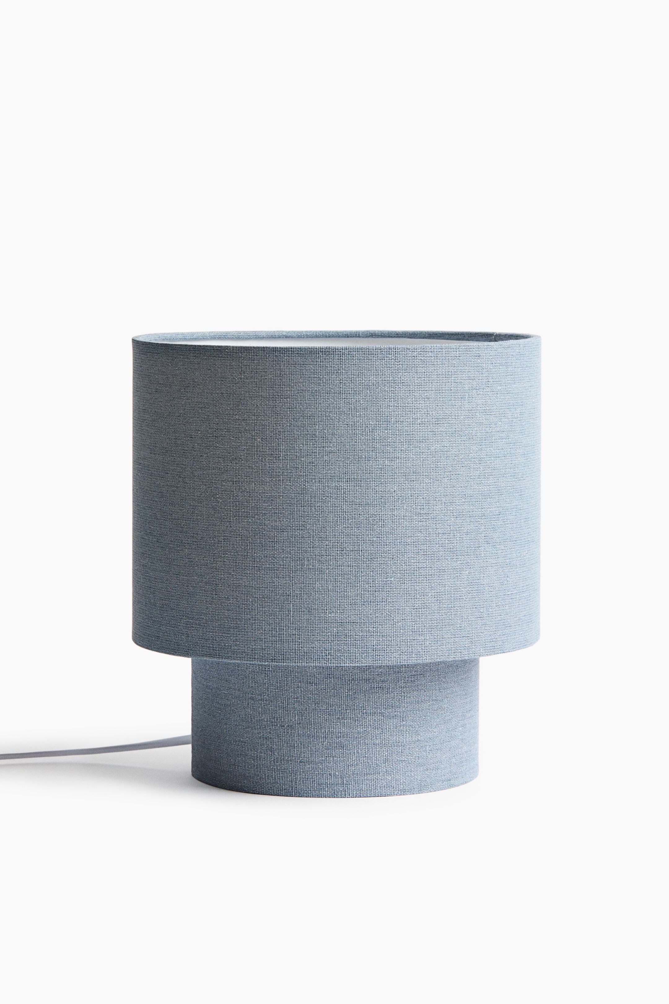 View larger image: A small table lamp stands front-facing, showcasing a cylindrical design made up of two stacked sections. The broader top section and the narrower base are enveloped in a light blue textured fabric, with a cord extending from the back.