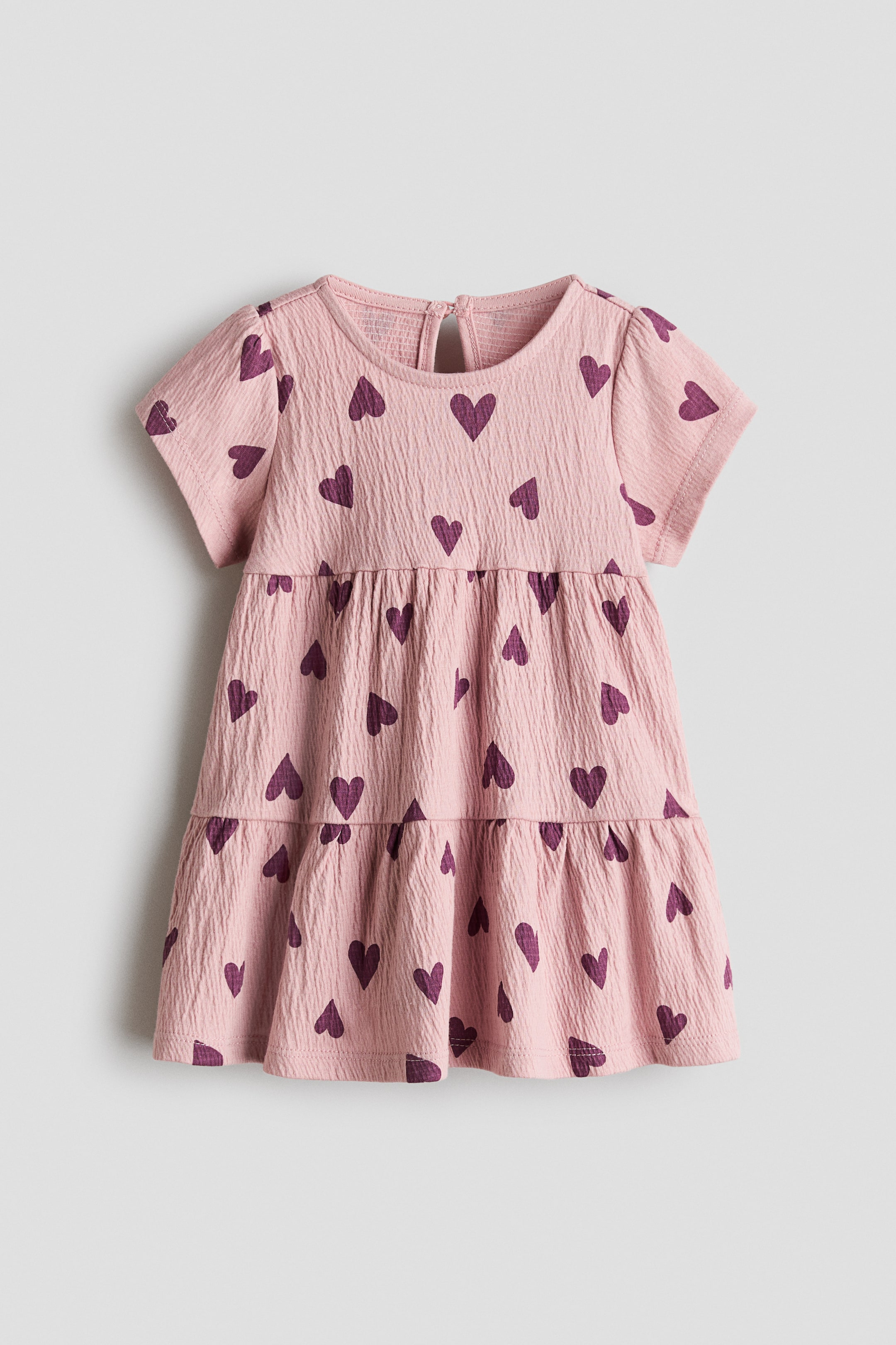 Xem ảnh lớn hơn: Chiếc váy cotton jersey màu hồng nhạt, có kết cấu, được đặt phẳng và hướng về phía trước, nổi bật với họa tiết trái tim màu tím đậm toàn bộ. Váy có cổ tròn, tay ngắn, lỗ khóa với nút ở phía sau, xếp ly ở eo và chân váy hai tầng xòe.