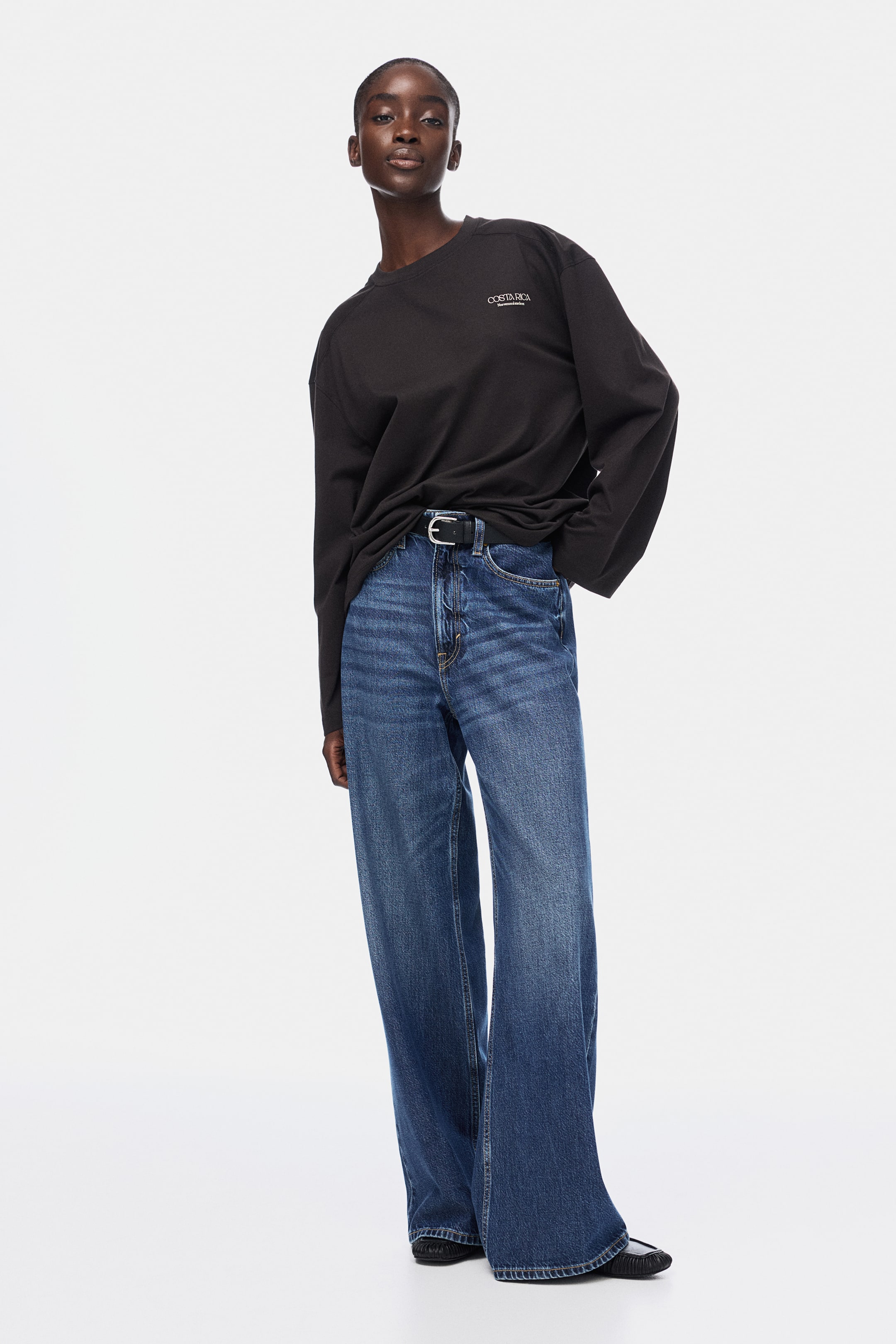 查看大圖: 一件黑色的宽松长袖T恤,配有圆领和左胸的白色文字,随意地塞进蓝色宽腿牛仔裤中。牛仔裤呈现出中等洗水效果,带有细微的猫须纹理,并搭配一条带银色扣的黑色腰带。