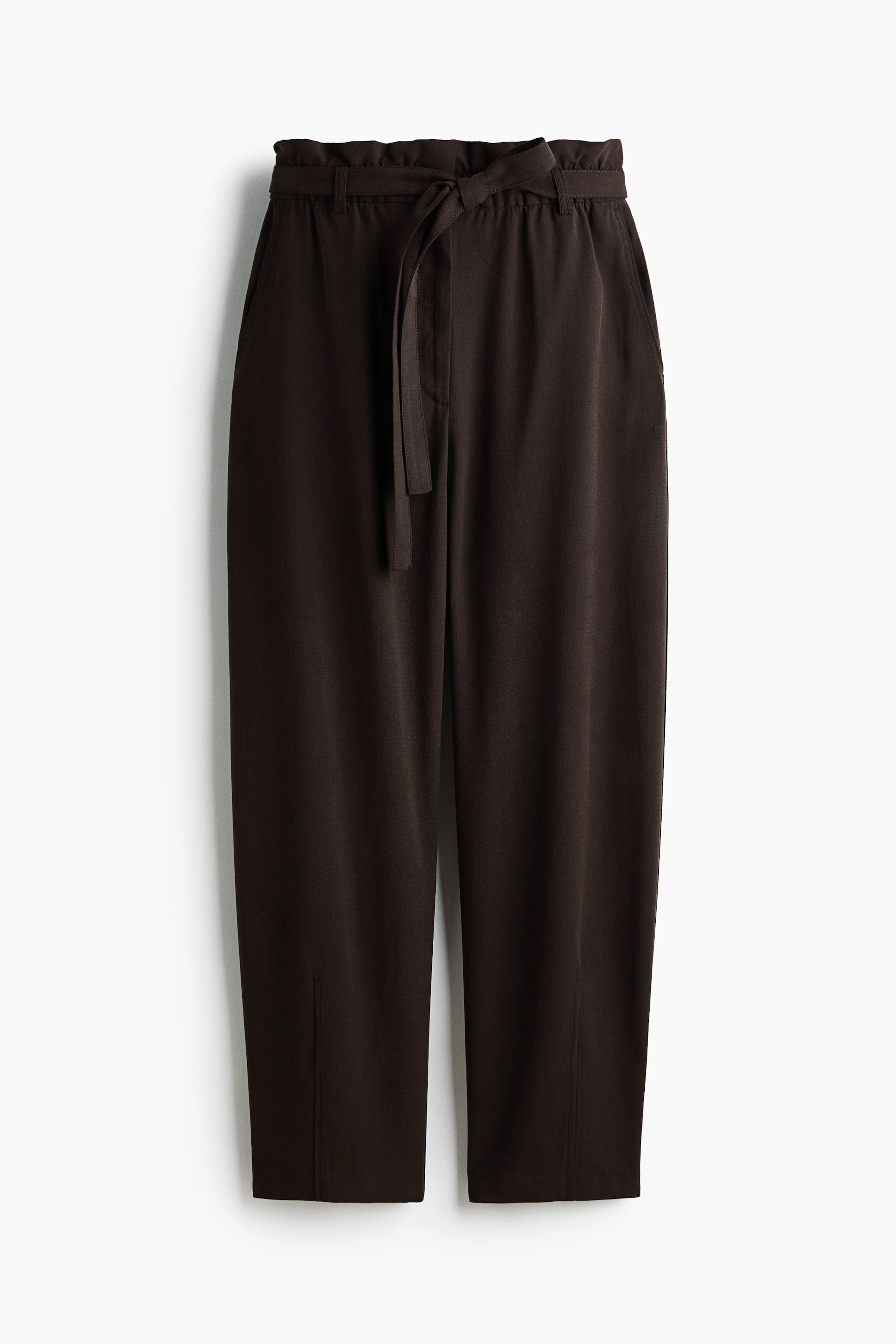 Ampliar la imagen: Pantalones oscuros de color marrón con una cintura alta y elástica tipo "paper bag", adornados con un cinturón ancho que se ata al frente. Estos pantalones a la altura del tobillo cuentan con discretos bolsillos laterales y piernas sueltas de corte barril con pinzas verticales en los dobladillos.
