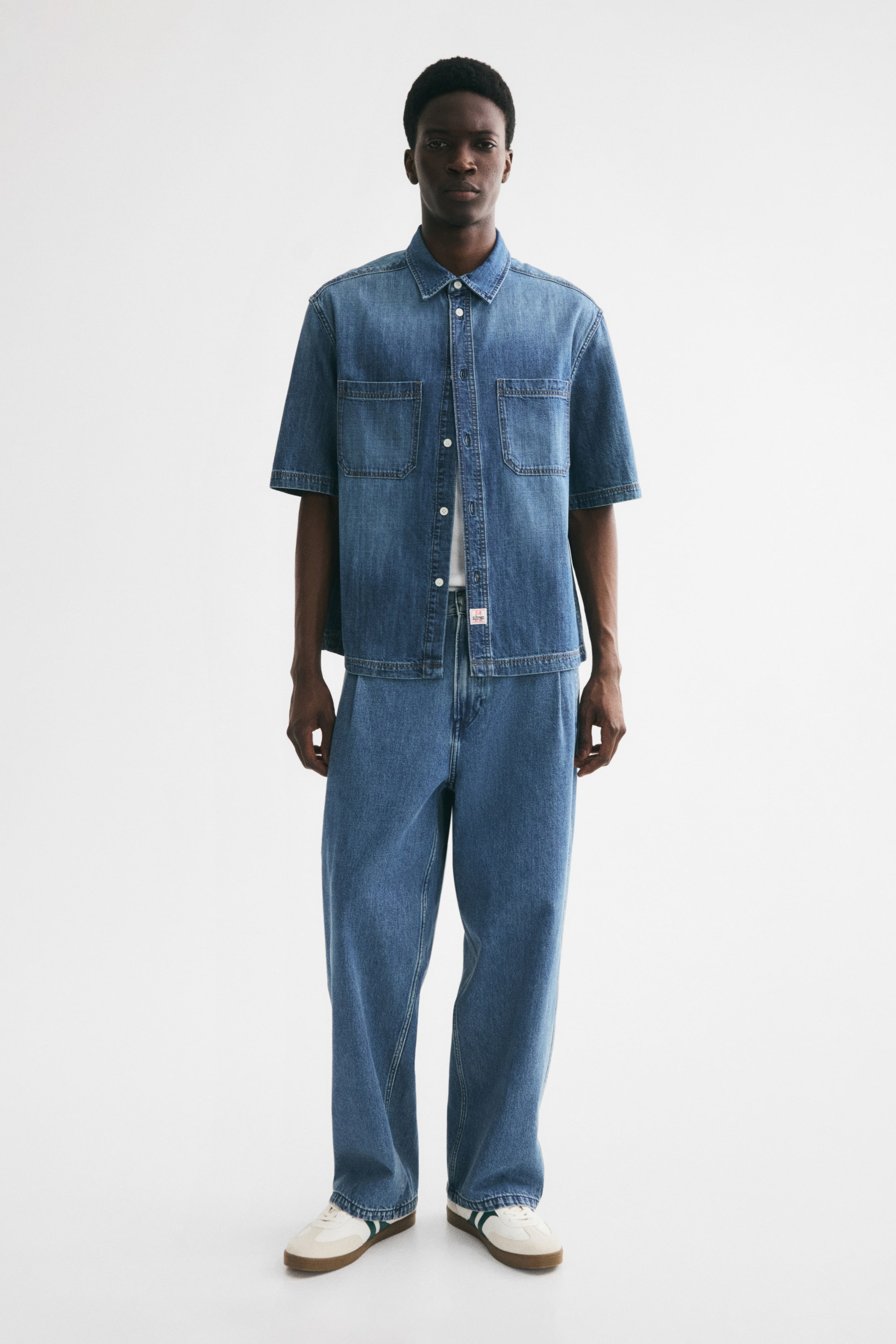 View larger image: The model is wearing jeans crafted from rigid cotton denim. These jeans feature a relaxed, baggy fit with a dropped crotch and ample room through the leg. They have a regular waist with a zip fly and button fastening, pleats at the top, side pockets, and straight legs.
