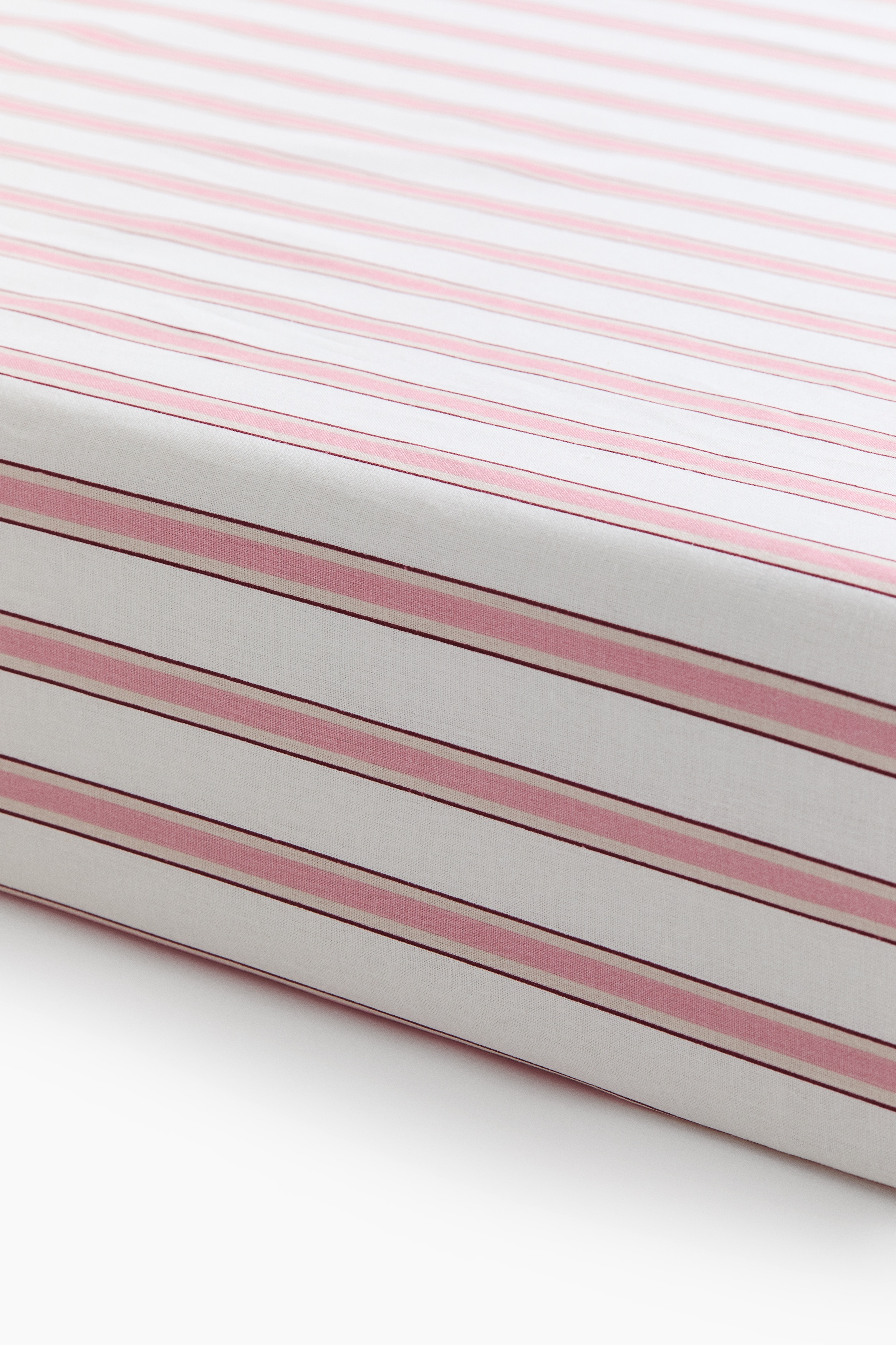 Un drap-housse en tissage de coton à motifs recouvre un matelas, affichant de fines rayures horizontales brun-rouge et blanches, avec un oreiller rose pâle posé dessus.