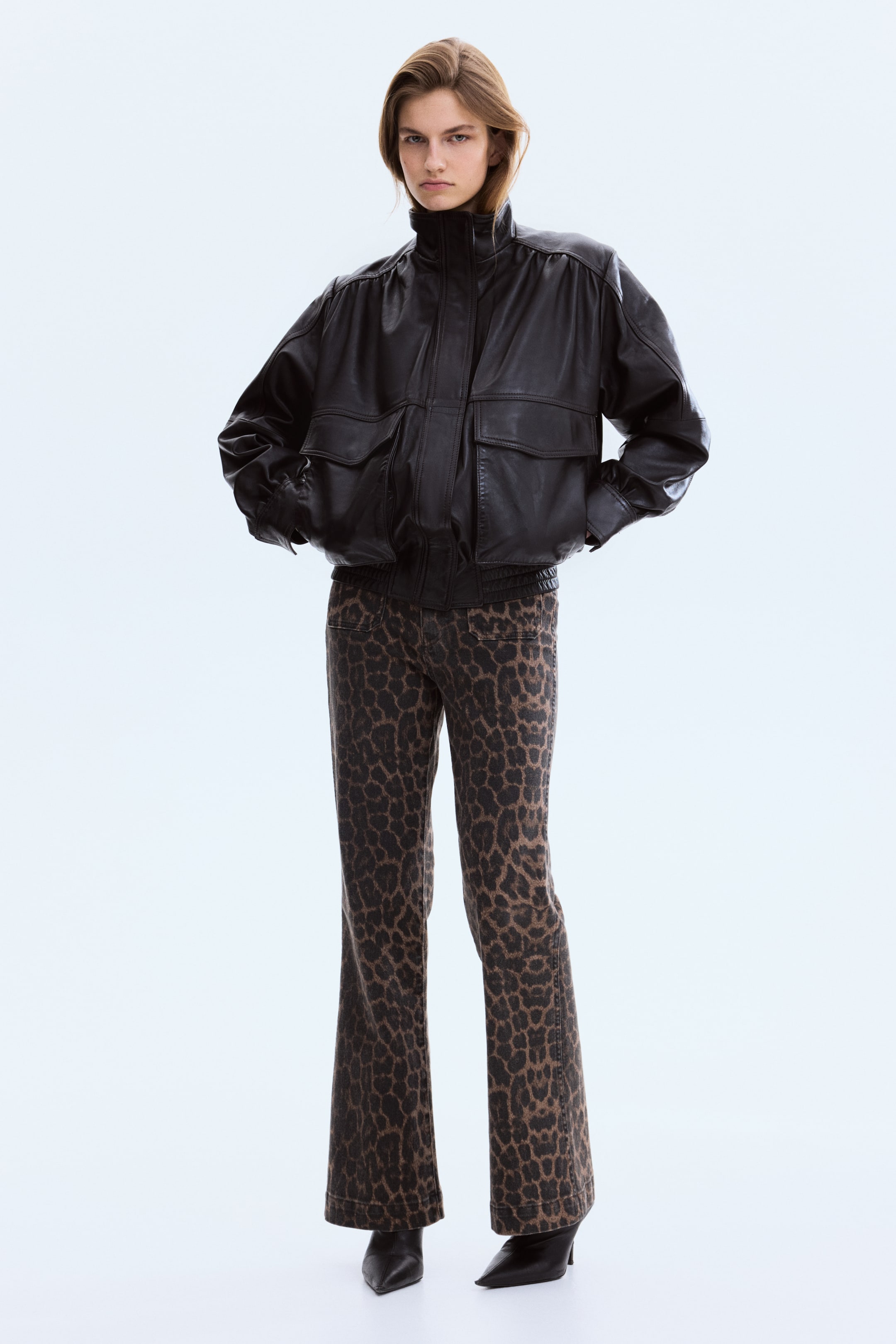 Visa större bild: En svart bomberjacka i läder med hög ståkrage, voluminösa rynkade ärmar med elastiska muddar, två stora fickor med lock framtill och en elastisk fåll, allt medan den är vänd framåt.