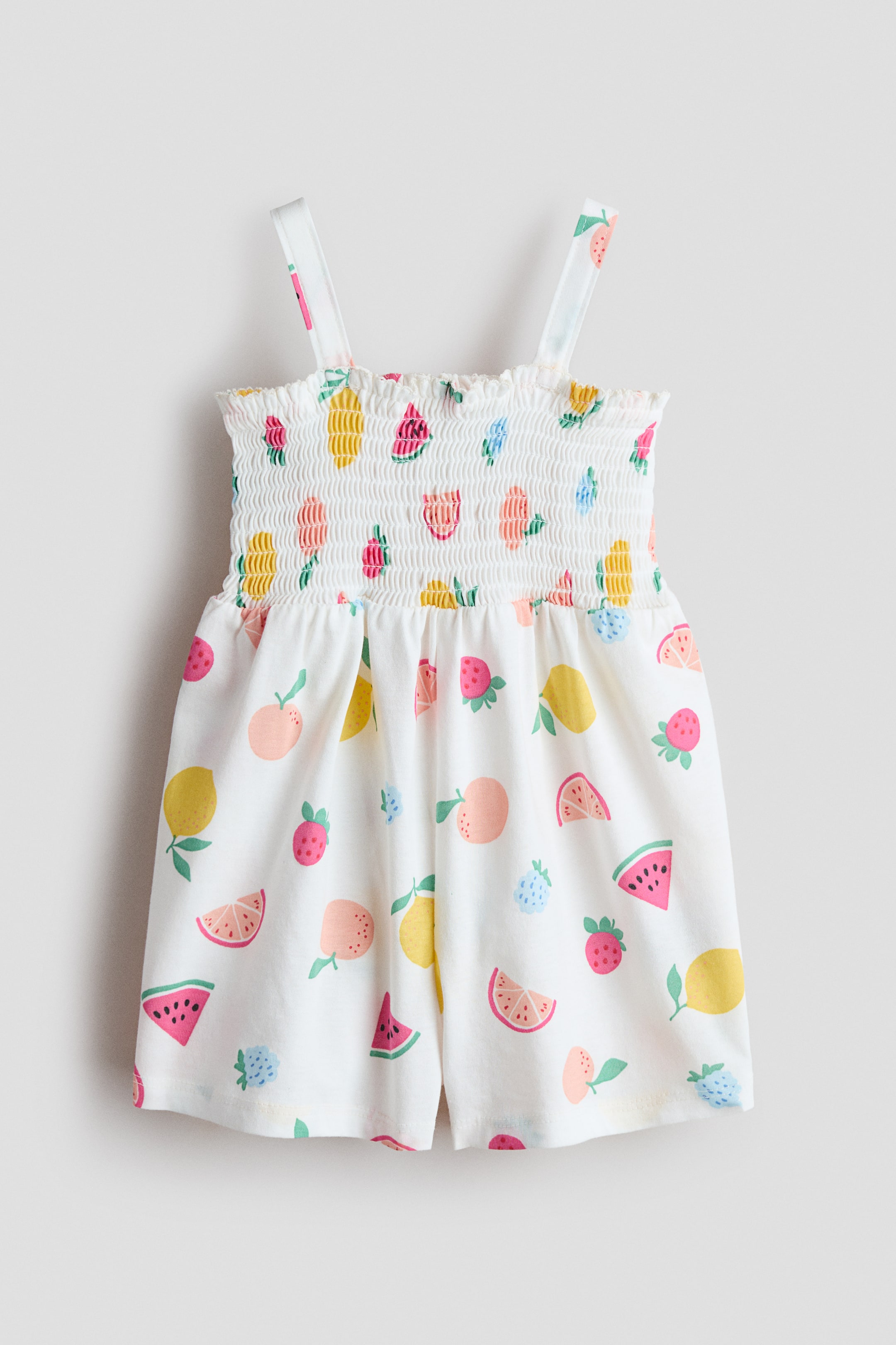 View larger image: The romper suit made of white cotton jersey showcases its front, adorned with a vibrant fruit pattern all over. It highlights smocking at the top, accented with a frill trim, a square neckline, slim shoulder straps, a gathered seam at the waist, and short legs.
