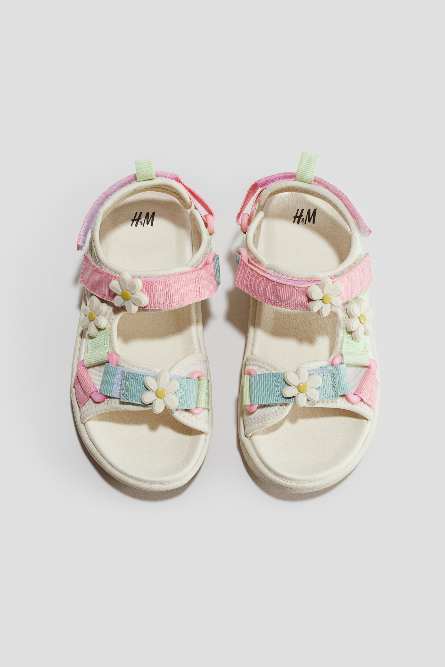 A pair of childrenâs scuba sandals is shown from the front in a top-down view, featuring off-white molded footbeds with adjustable pastel pink, light blue, and light green hook-and-loop straps. The straps are decorated with small, three-dimensional white daisy flower appliquÃ©s with yellow centres.