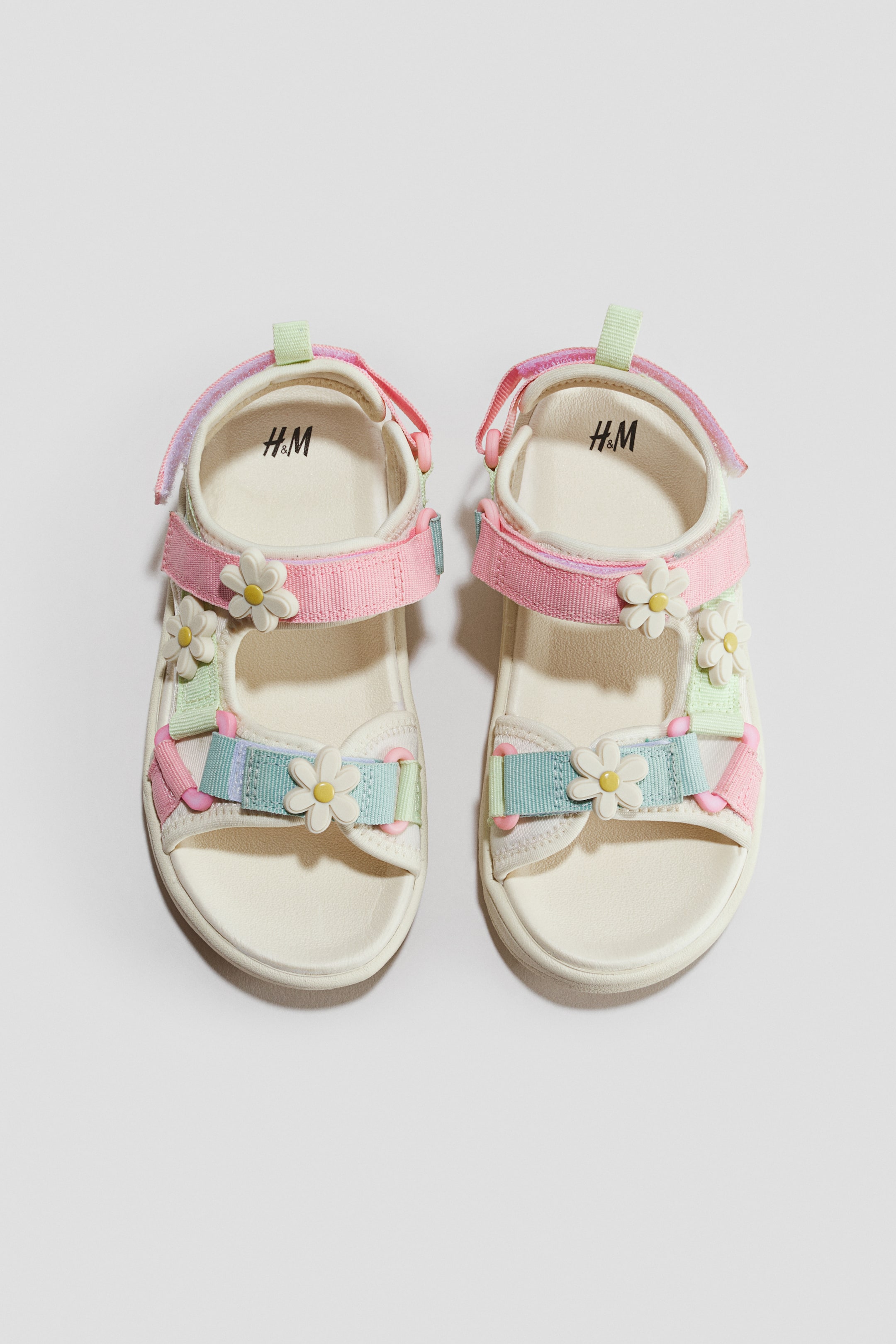 View larger image: A pair of children’s scuba sandals is shown from the front in a top-down view, featuring off-white molded footbeds with adjustable pastel pink, light blue, and light green hook-and-loop straps. The straps are decorated with small, three-dimensional white daisy flower appliqués with yellow centres.