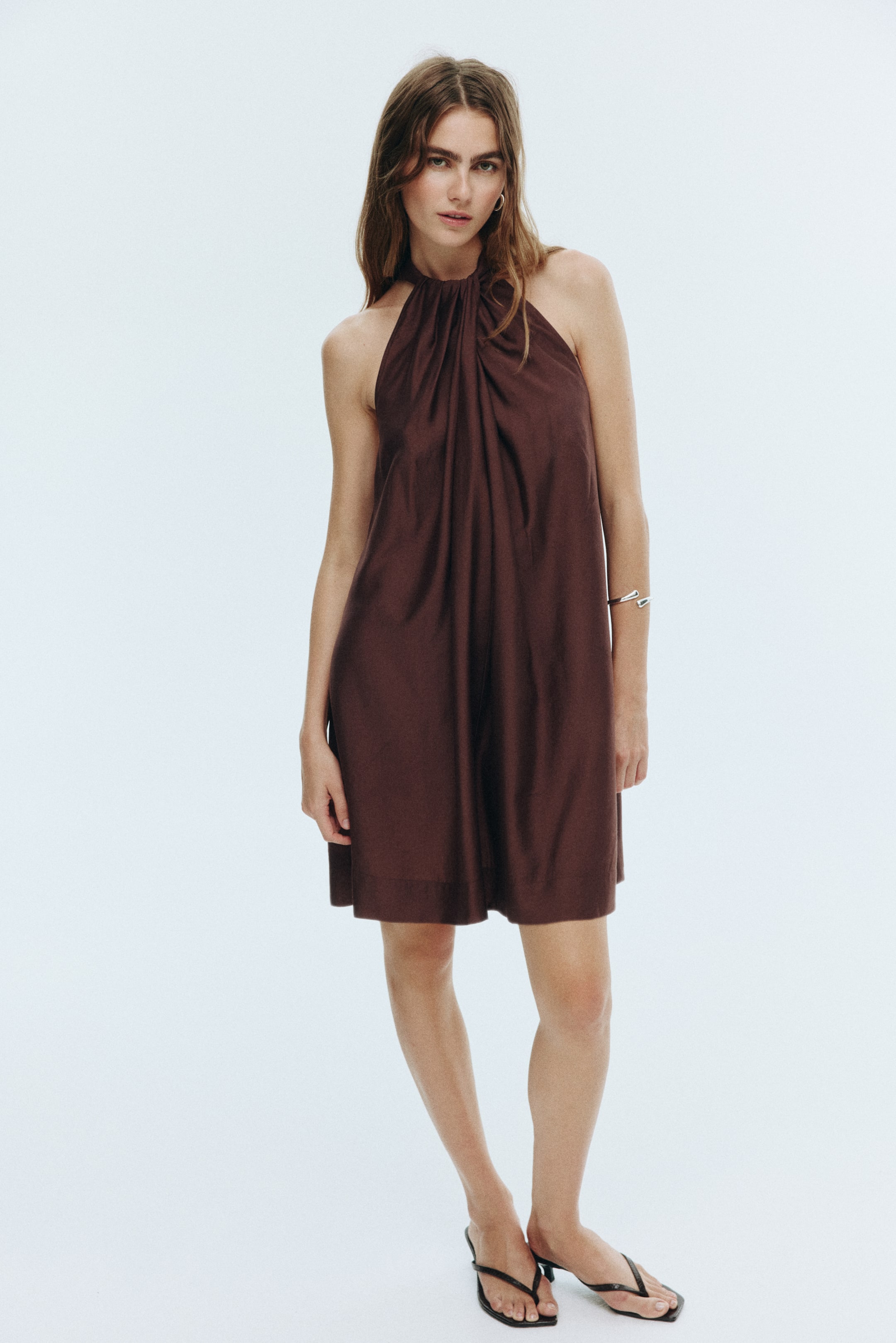 View larger image: A deep brown halter neck dress is being displayed, facing forward. The dress is featuring a gathered and twisted detail at the neckline, which is flowing into a relaxed, A-line silhouette that is ending above the knee.