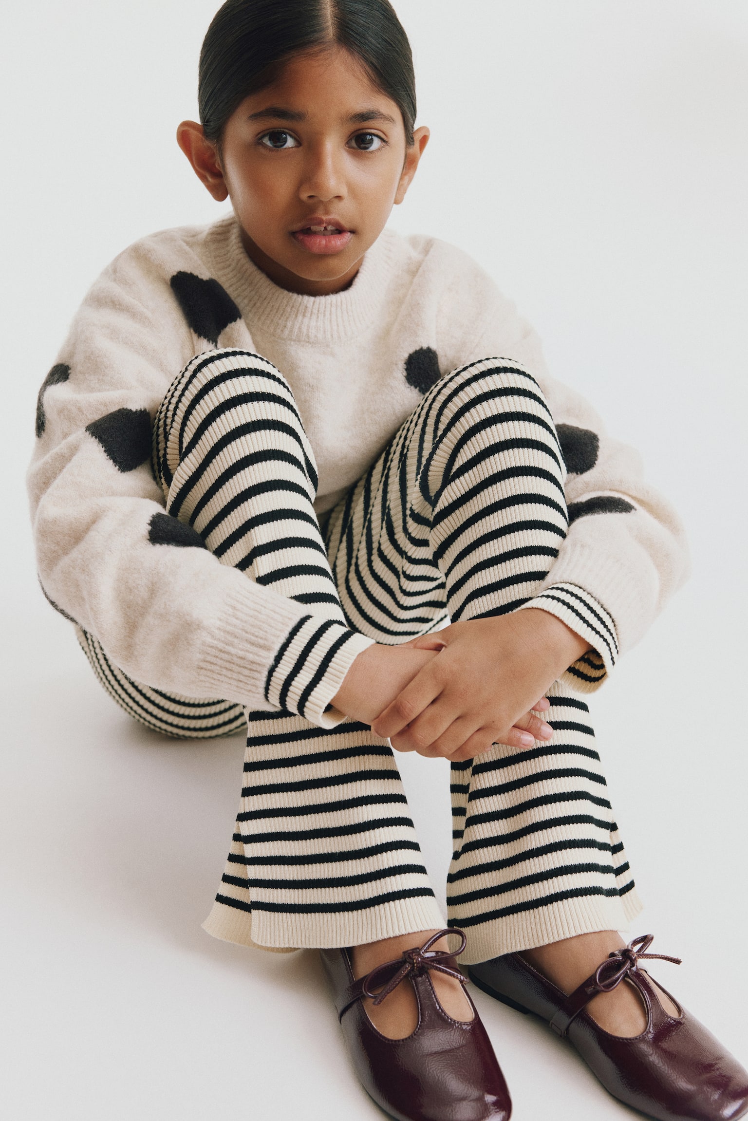 Striped rib-knit leggings in cream and black are being worn by a seated model facing forward, highlighting their fitted silhouette and horizontal pattern. The leggings flare at the hem, showcasing a side slit at the outer ankle. - 1