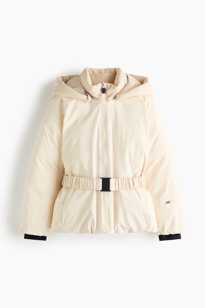 Eine cremefarbene Steppjacke ist von vorne zu sehen und verfügt über eine angesetzte Kapuze, einen hohen Kragen und einen mittigen ReiÃverschluss. Sie zeigt einen elastischen Taillengürtel mit schwarzer Schnalle, schwarze Innenbündchen und eine kleine ReiÃverschlusstasche am linken Ãrmel. - 1