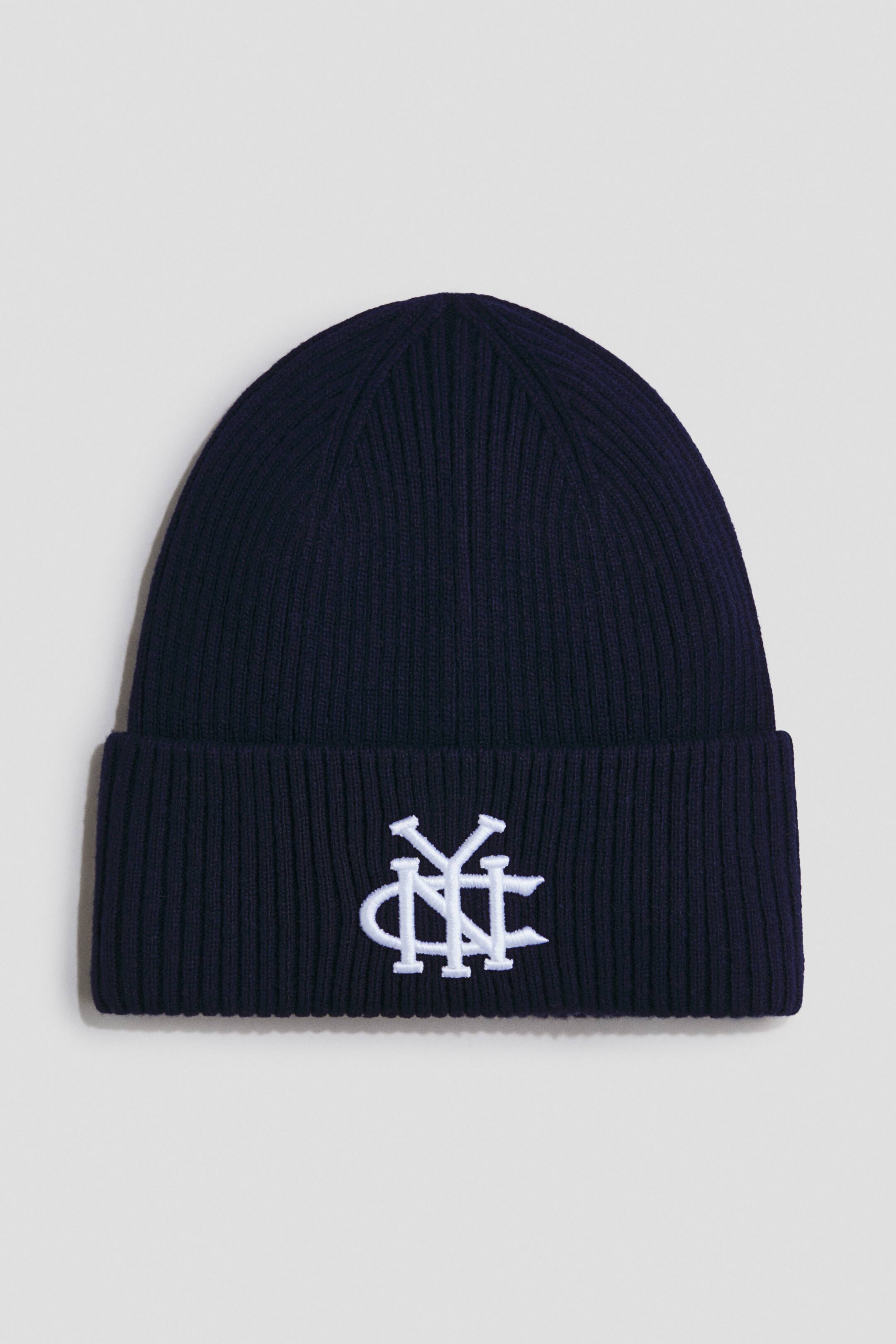 Grotere afbeelding bekijken: Een marineblauwe ribgebreide beanie heeft een omgeslagen boord met op de voorkant een wit geborduurd gestileerd monogram.