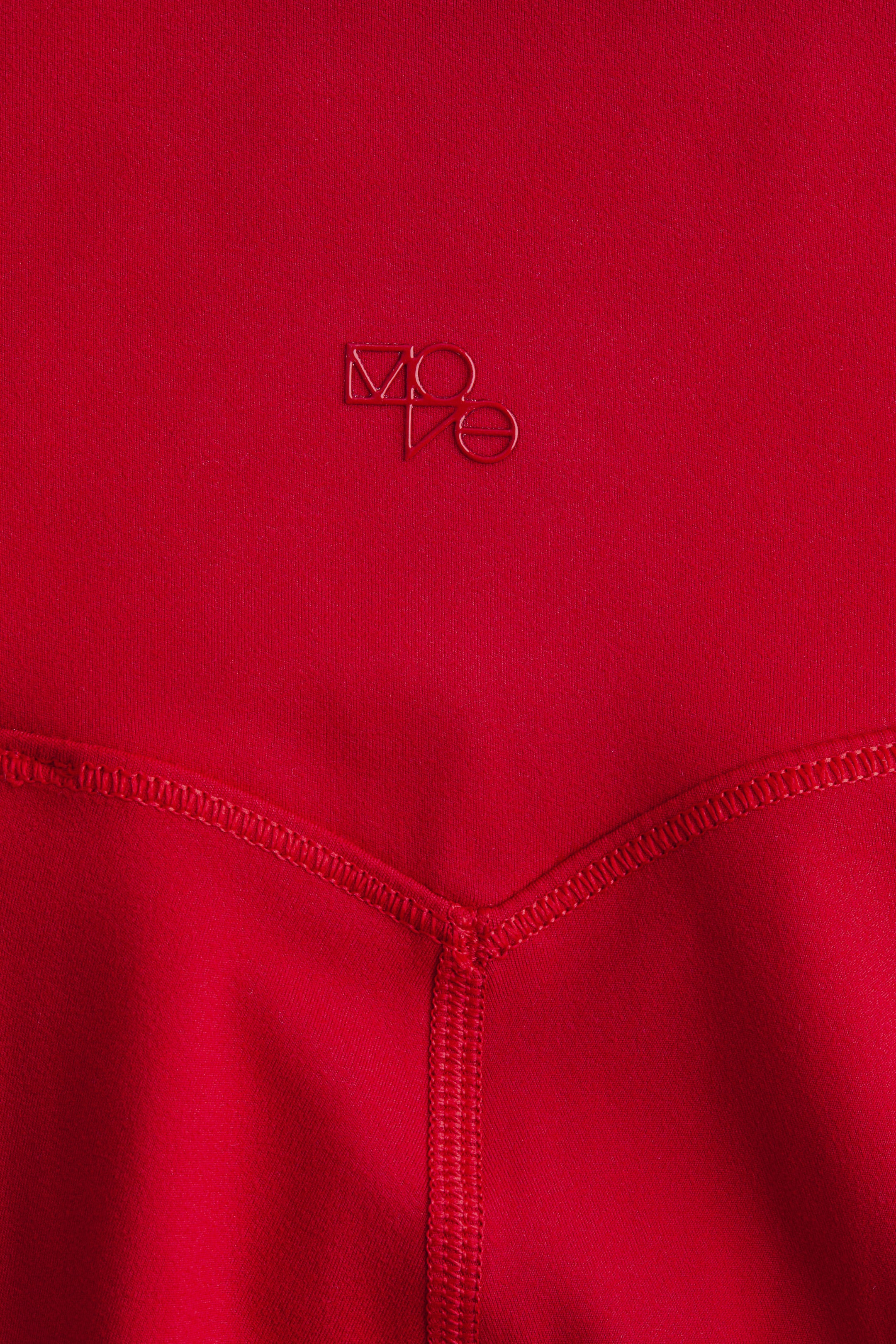 View larger image: Red sports leggings with SoftMove? are featuring a stylized, embossed red 'MOVO' logo on the upper front section. Prominent V-shaped flatlock stitching is appearing below the logo, extending vertically downwards, suggesting a structural seam.