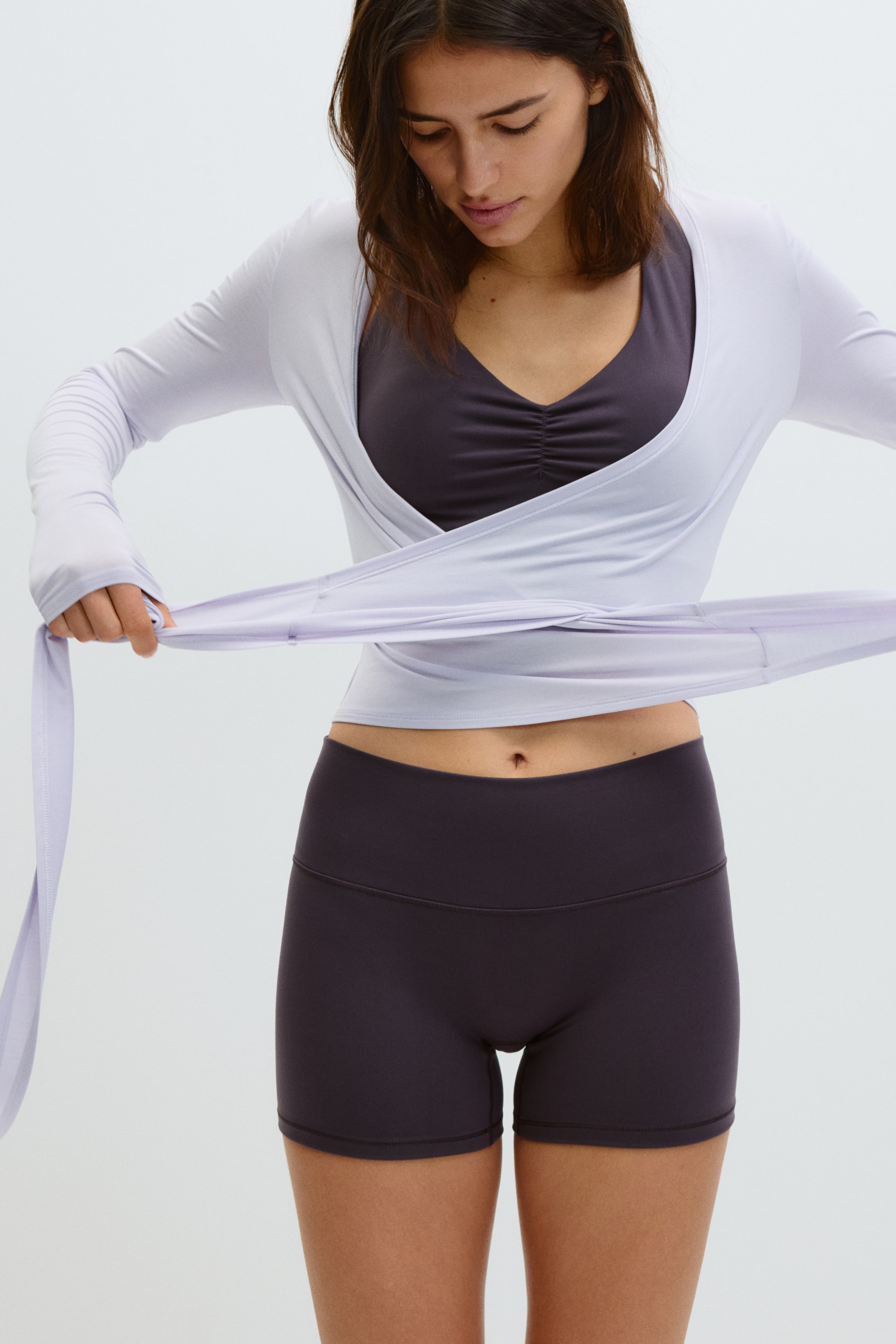 Ver imagen más grande: Un top cruzado de manga larga en color lila claro se cruza sobre el pecho y se ata alrededor de la cintura, mostrado desde un ángulo frontal. Las mangas largas con agujeros para los pulgares se extienden desde el top, que se superpone a un sujetador deportivo gris oscuro, visible a través de su escote en V.