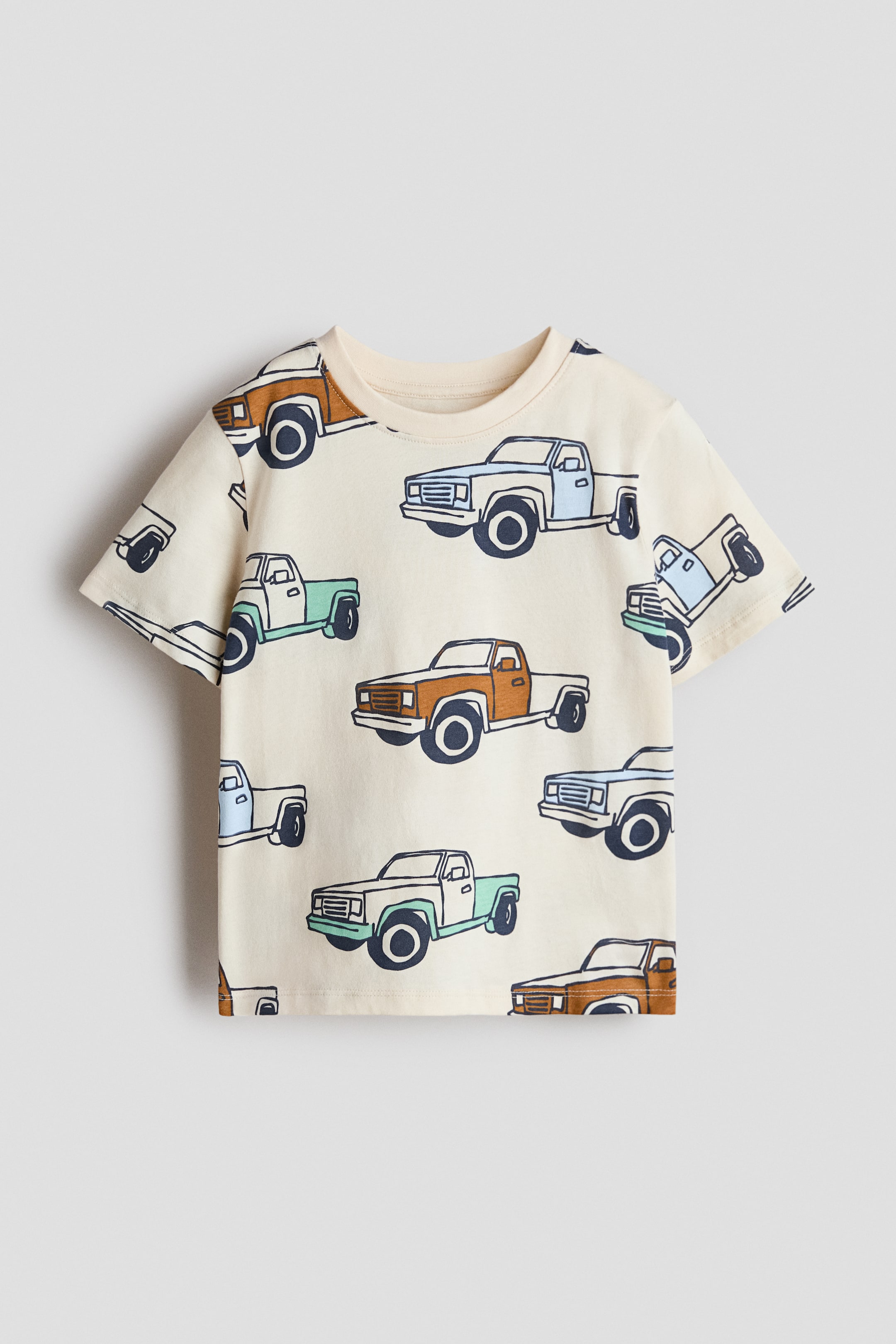 View larger image: The T-shirt is shown FRONT, featuring a round neckline and short sleeves. It is made of soft, printed cotton jersey, displaying an all-over pattern of light blue, green, and brown pickup trucks on a cream background.