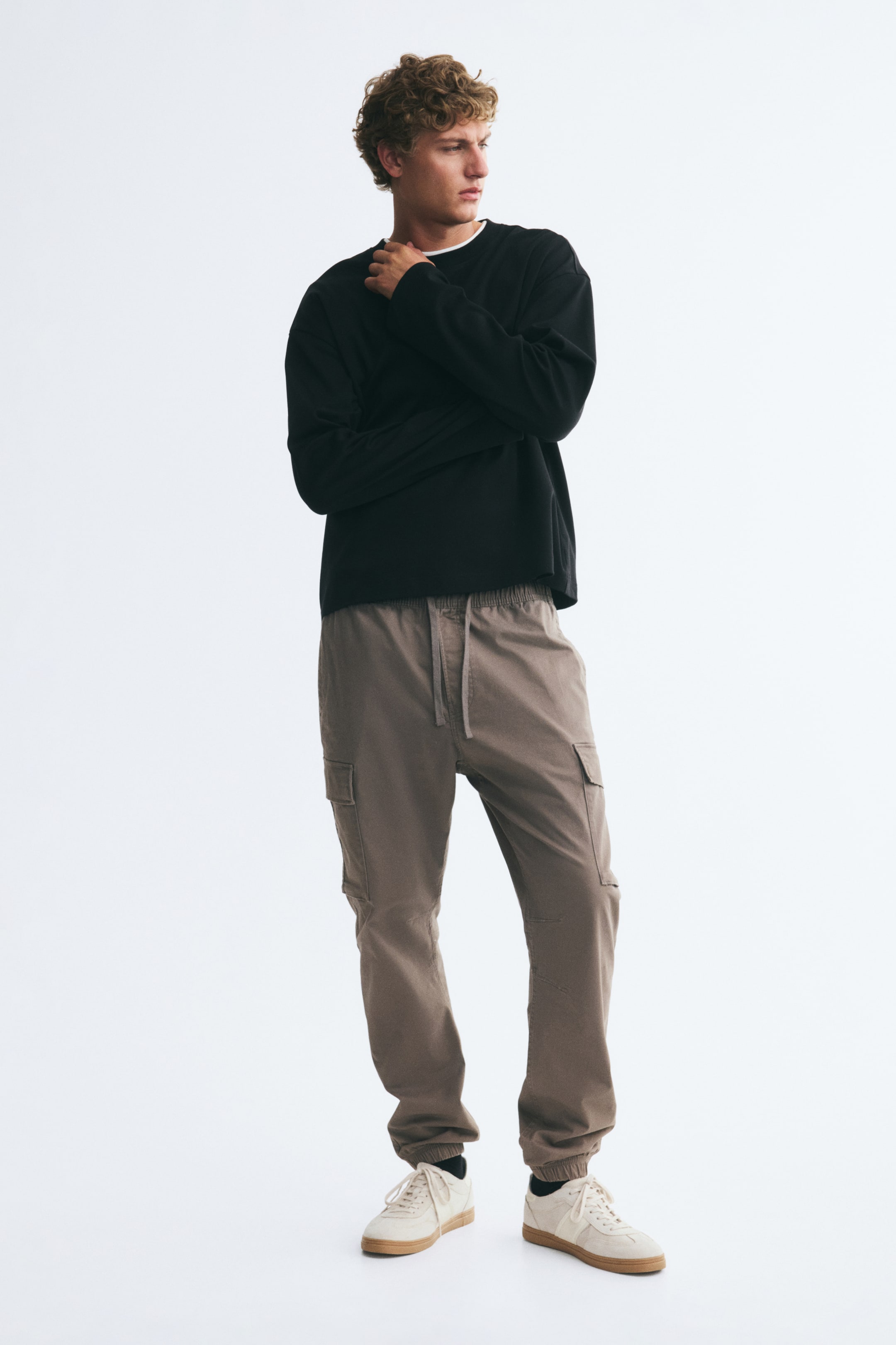 View larger image: Taupe cotton twill trousers featuring an elasticated drawstring waist, cargo flap pockets at the sides, and tapered legs with elasticated hems are worn by the model. The trousers are styled with a black long-sleeve sweatshirt and off-white trainers.
