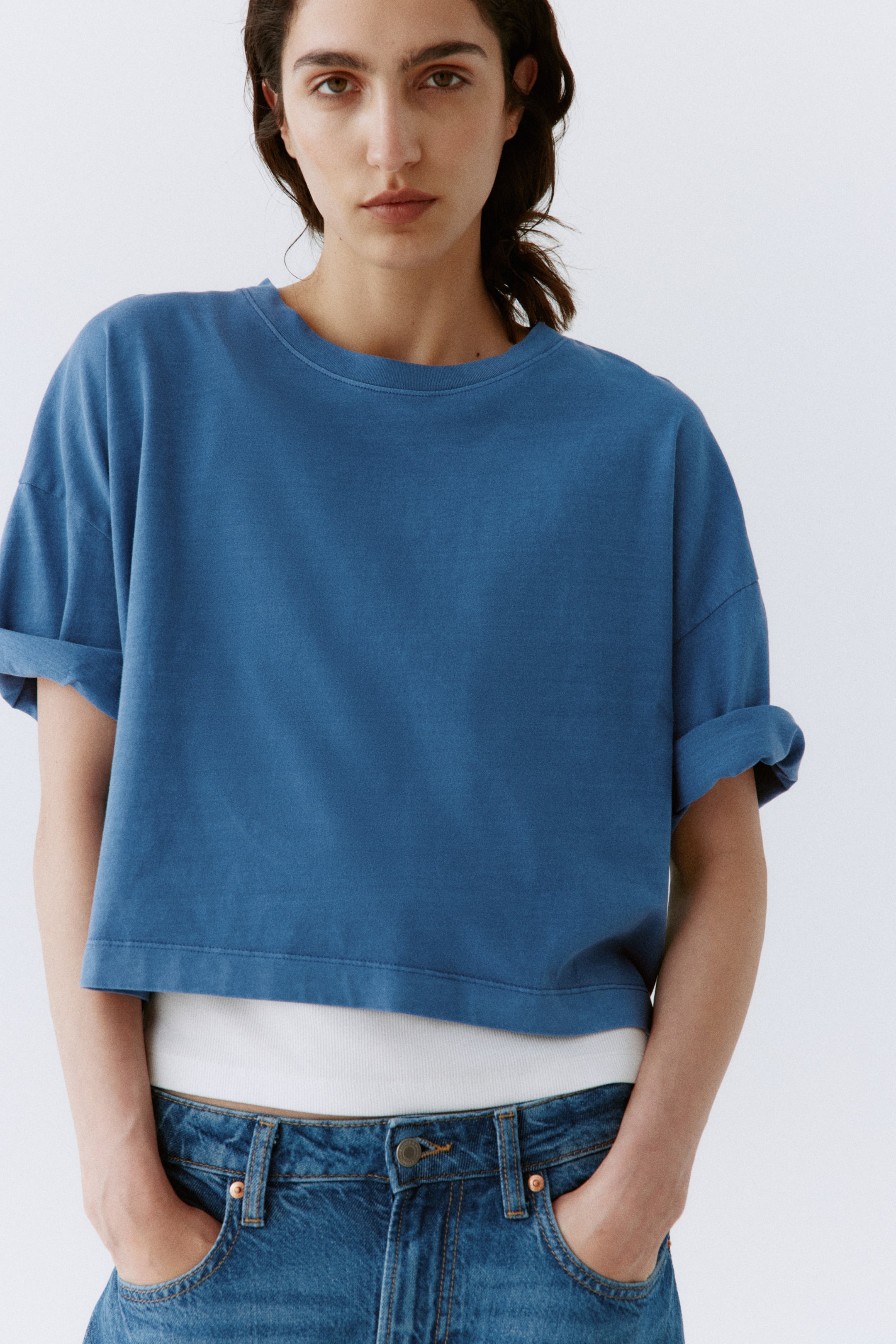 Visualizza immagine più grande: Una t-shirt corta blu presenta un girocollo e maniche corte arrotolate, offrendo una vestibilità comoda e squadrata. L'orlo dritto e corto rivela una maglietta bianca sottostante.