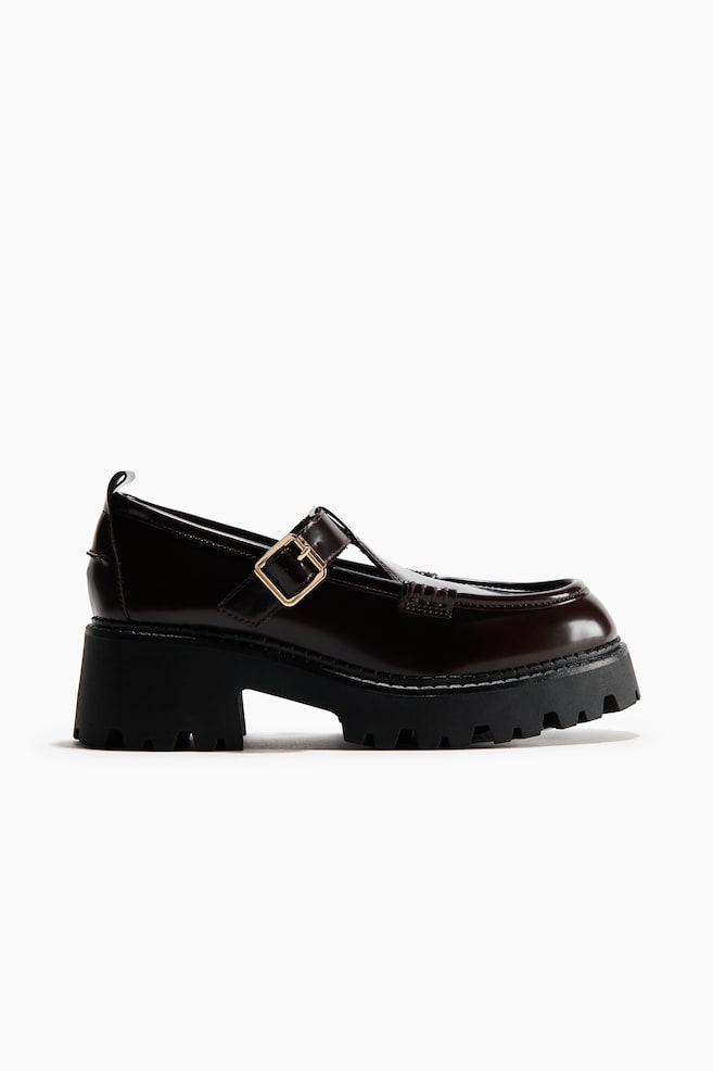 A dark brown, glossy Mary Jane shoe is showcasing its side profile, featuring a gold-toned buckle on a strap across the instep, a rounded moc toe, and a thick black lug sole with a chunky block heel. - 1
