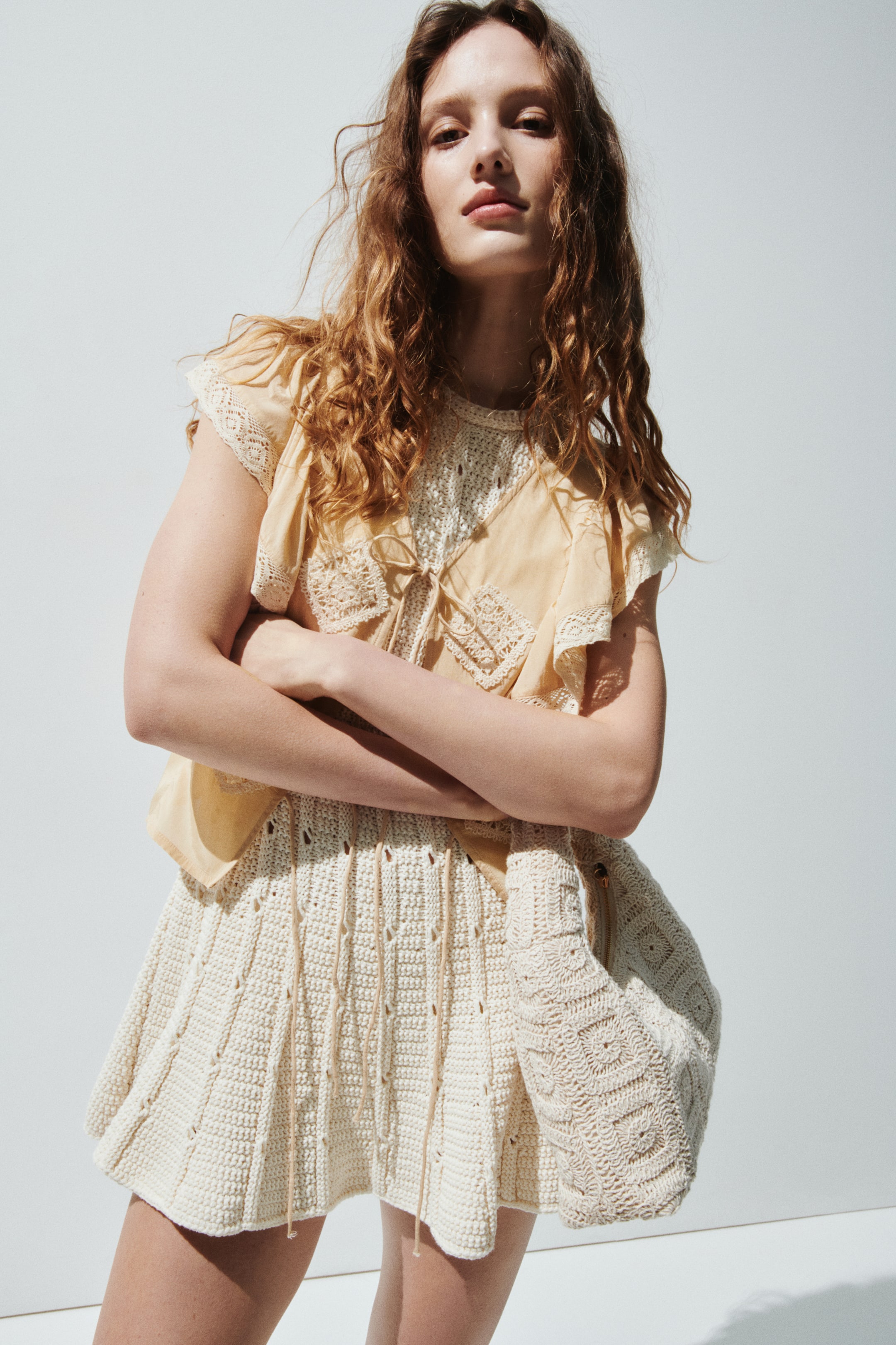 View larger image: The outfit showcases a pale yellow short-sleeved top adorned with ruffled, lace-trimmed sleeves and a V-neck with ties, layered over a cream crocheted high-neck top. Complementing this is a matching cream crocheted A-line skirt with vertical openwork details, and a coordinating crocheted bag featuring square patterns is carried at the side.