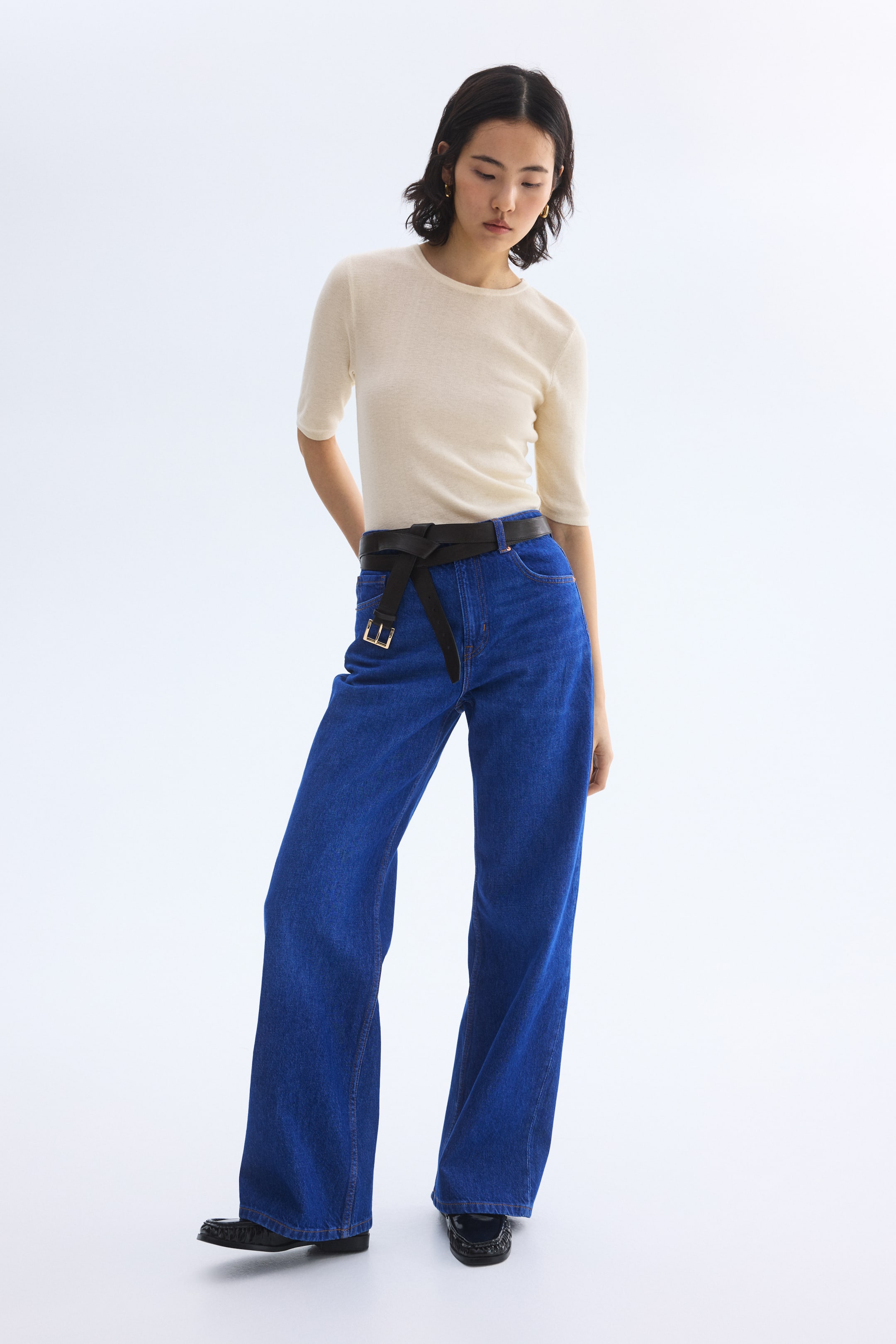 View larger image: A cream-coloured, close-fitting, crew neck top with elbow-length sleeves is being worn, tucked into vibrant blue high-waisted wide-leg jeans. A black belt with a gold-tone buckle is being tied around the jeans' waist, and black loafers with a textured vamp are visible.