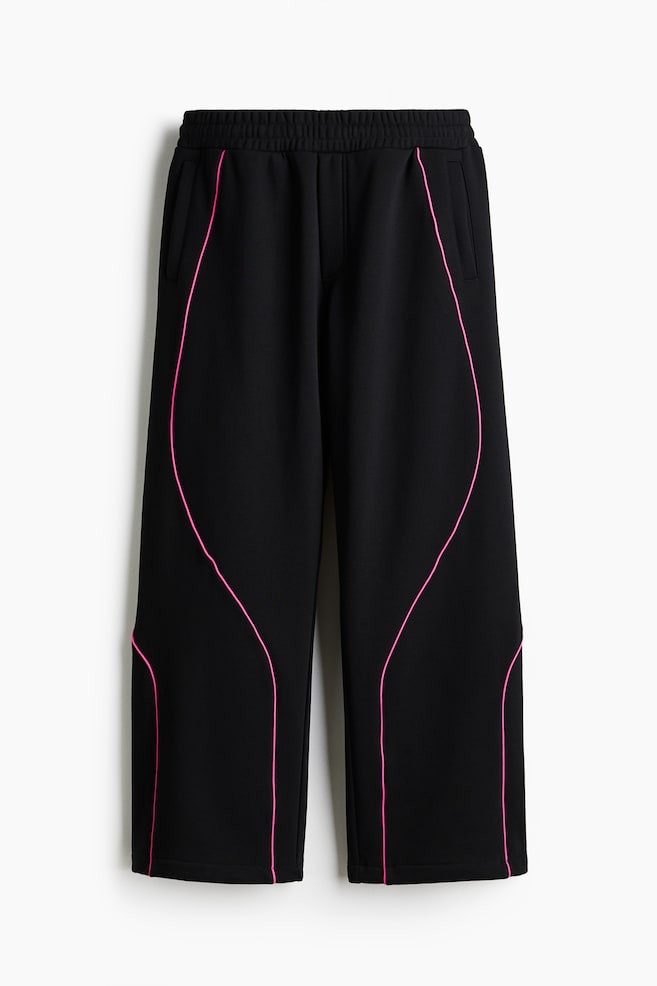 Schwarze Hosen werden von vorne präsentiert, mit einem elastischen Bund und Seitentaschen. Geschwungene rosa Paspeln zeichnen prominent ein wellenförmiges Muster entlang der Vorderseite jedes Beins nach. - 1