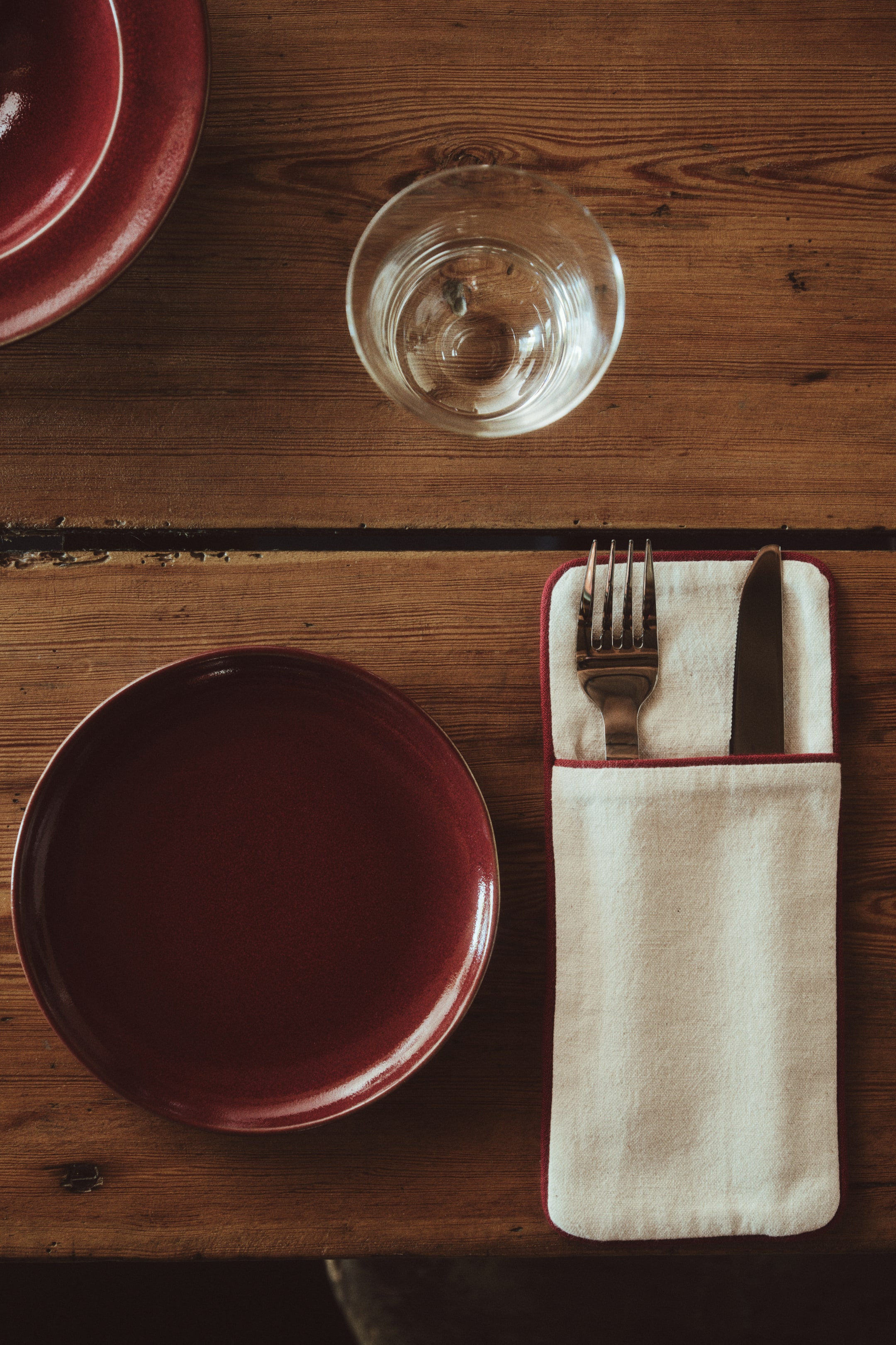 View larger image: A cutlery pouch in an off-white cotton and linen weave is shown from a top-down view, featuring dark red accent piping along the edges. The pouch is holding a metal fork and knife inside its front pocket.