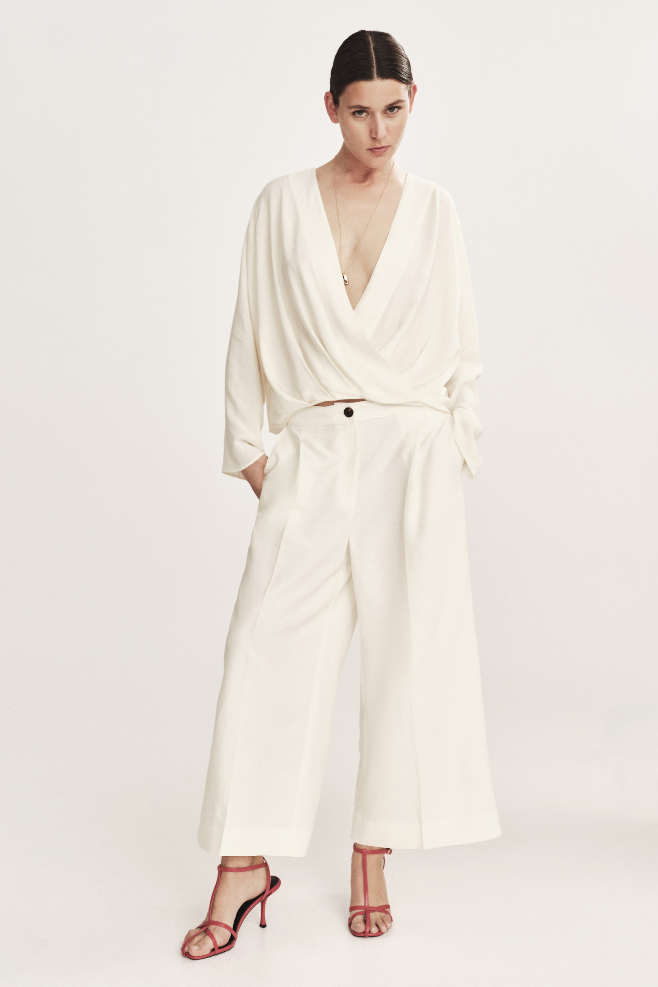 View larger image: A front-facing off-white matching set is showcased, featuring a draped, wrap-style blouse with a deep V-neck, accessorized with a gold pendant necklace, and wide-leg, pleated cropped pants. The pants are fastened by a black button and paired with red strappy high-heeled sandals.