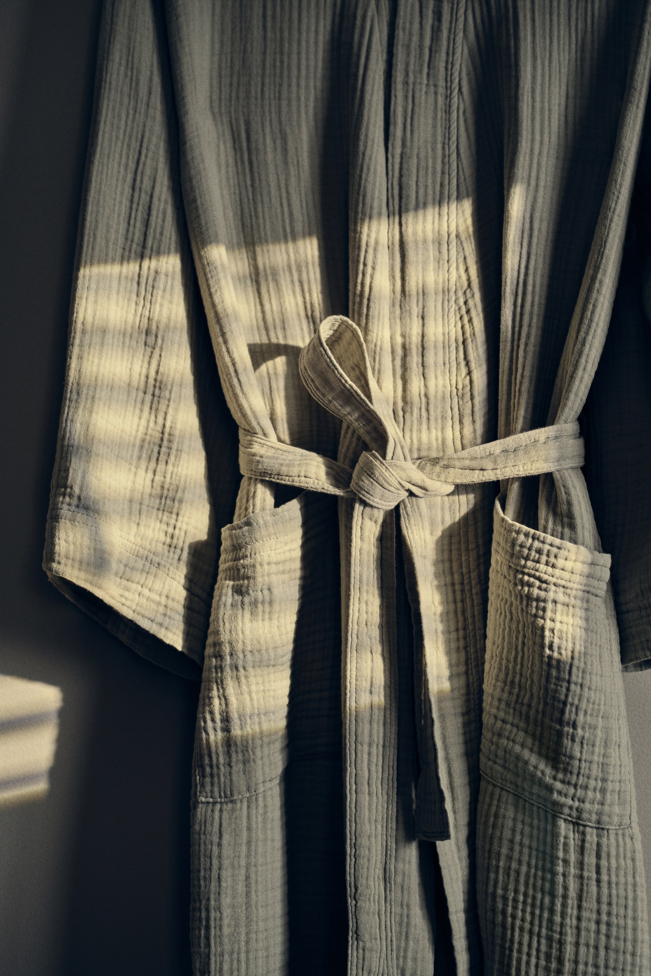 Se større billede: En lysegrøn salvie farvet morgenkåbe hænger, lavet af krøllet gaze stof. Den har et bælte af samme stof bundet i taljen og to store påsyede lommer foran.