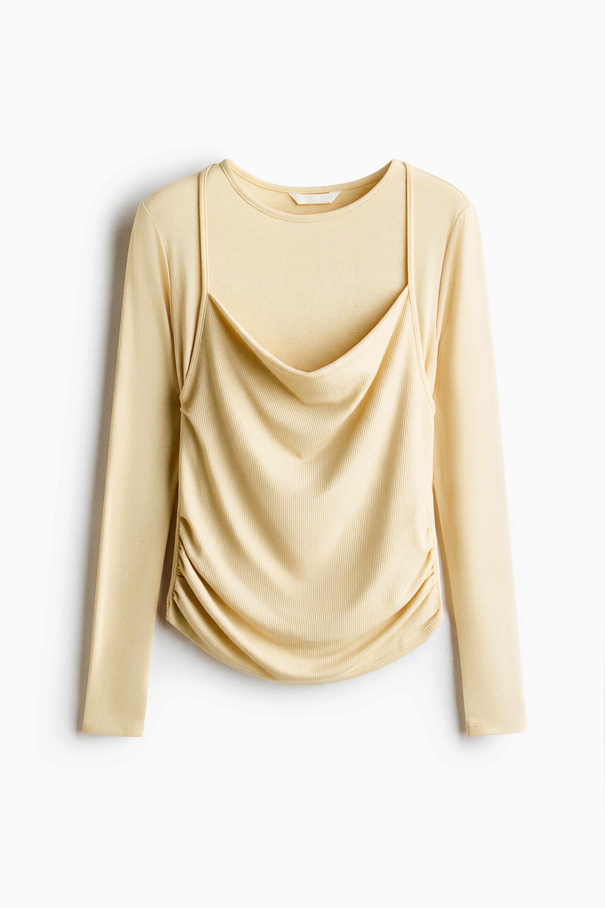 A light yellow, long-sleeved jersey top is worn by the model, featuring a high round neckline and a layered, cami-style front panel with a draped cowl neck and thin halterneck ties. The top has gathered side seams that create a draped effect at the waist and is paired with a dark green pleated skirt, black tights, and black loafers.
