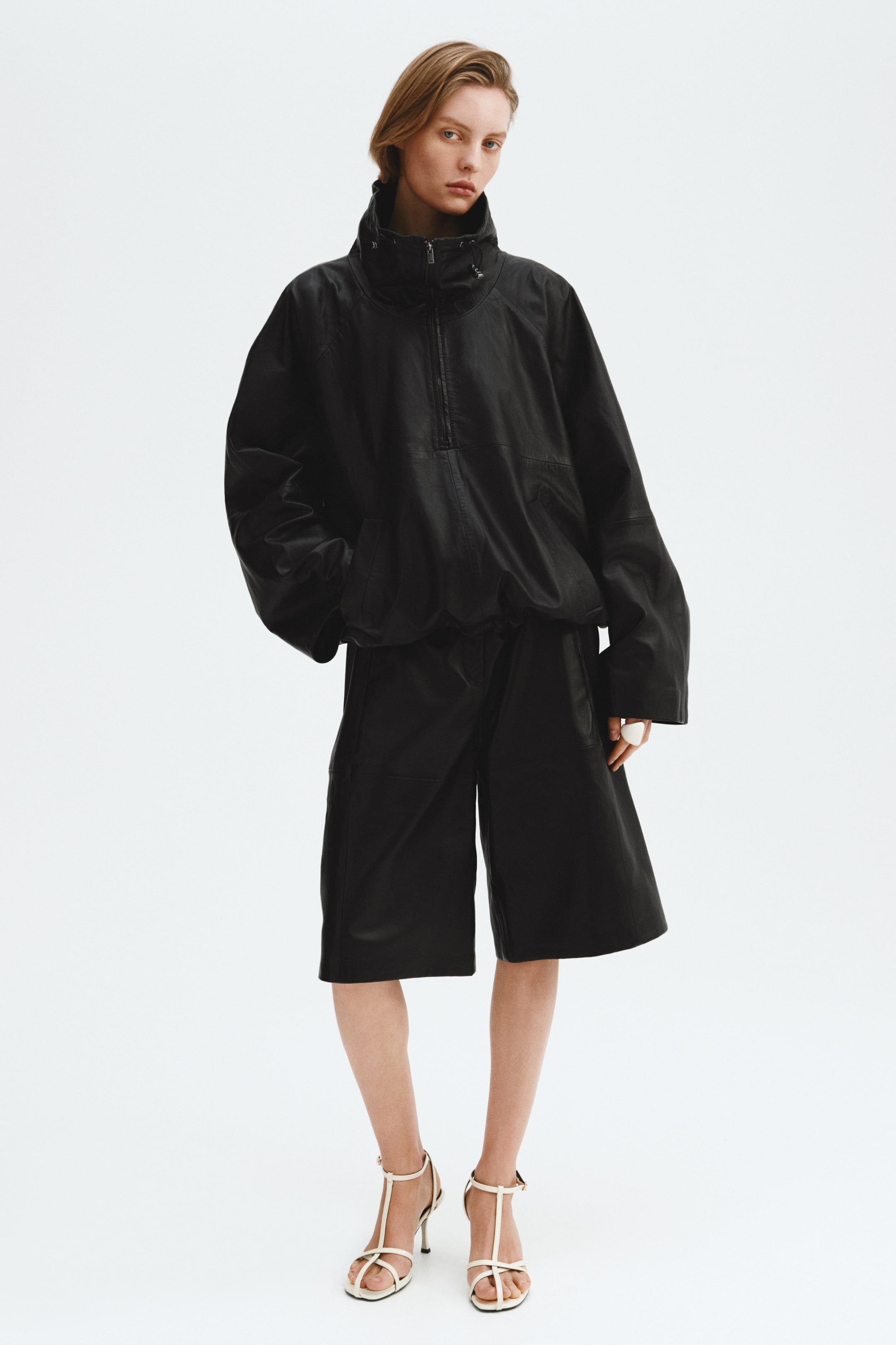 View larger image: A black leather anorak with a high stand-up collar, visible drawstring, and half-zip front closure is showcased on a model facing forward. The anorak also features wide, voluminous sleeves and is paired with matching knee-length shorts that exhibit a relaxed fit.