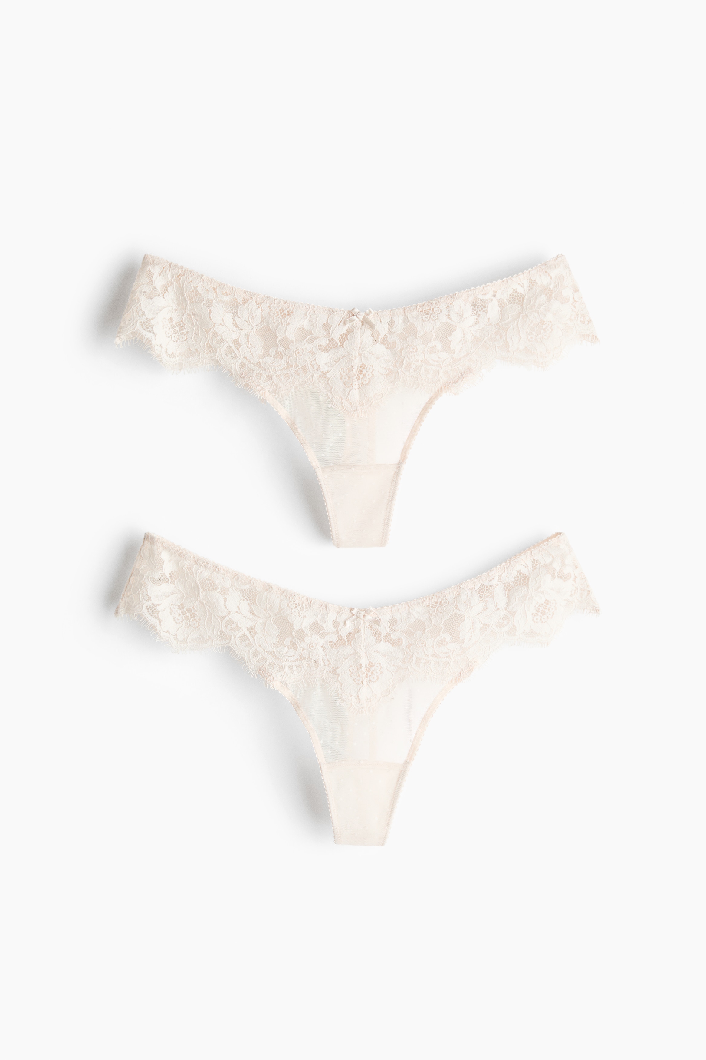Een witte kanten string met lage taille en geschulpte randen wordt gedragen door het model, dat in zijprofiel staat. Het model draagt ook een bijpassende witte kanten en mesh bh.