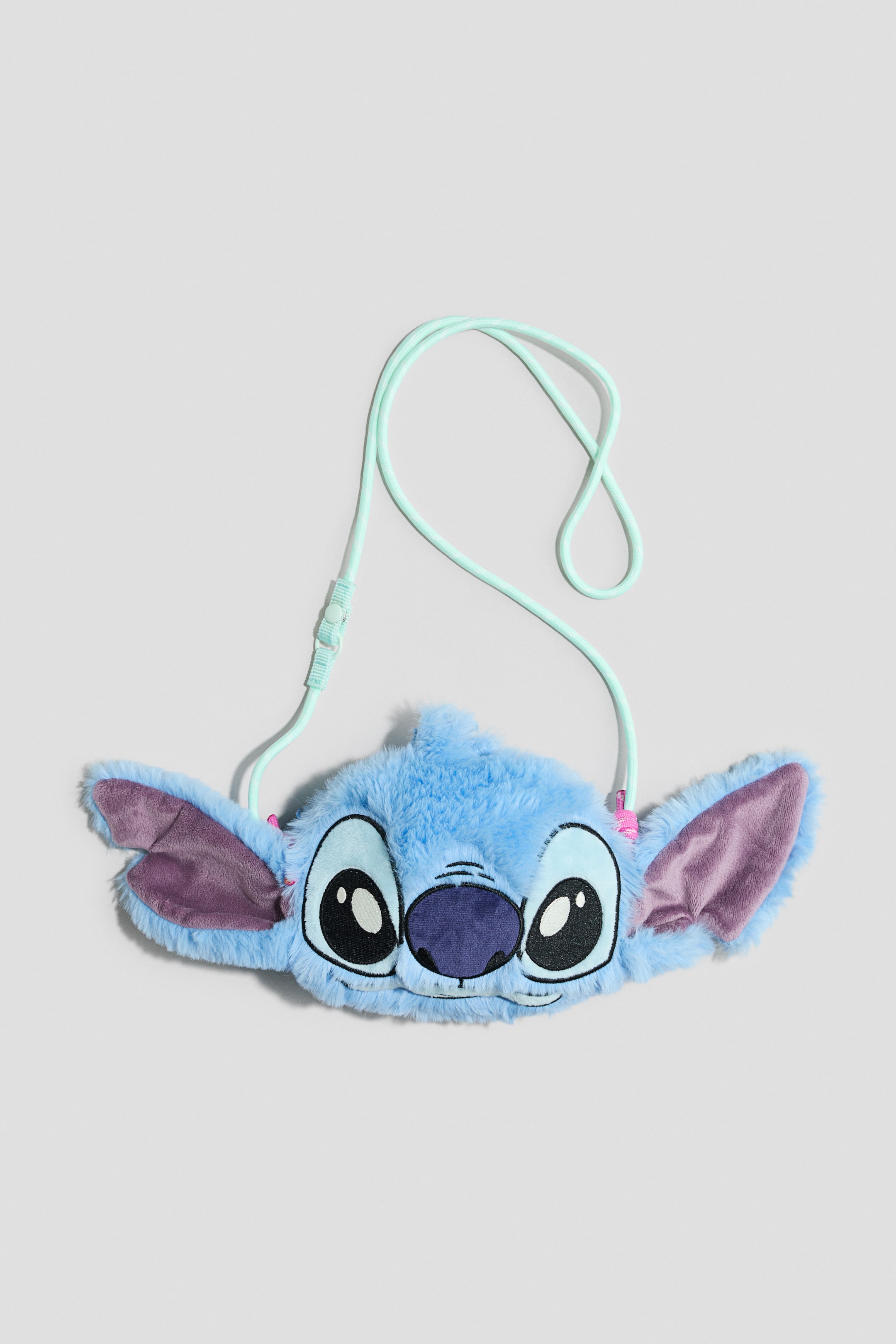 Näytä suurempi kuva: Sumea sininen Stitch-muotoinen pussukka makaa litteänä, esitellen kirjailtuja kasvonpiirteitään, mukaan lukien suuret mustavalkoiset silmät, tummanvioletti nenä ja hienovaraiset valkoiset hampaat. Siinä on suuret violettireunaiset korvat ja ohut, vaaleanvihreä säädettävä nyöriremmi.