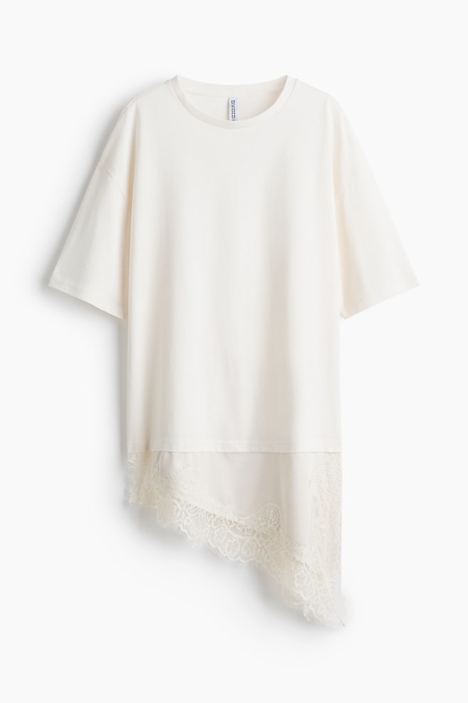 Een off-white t-shirt met een ronde hals en korte, wijde mouwen, voorzien van een asymmetrisch crèmekleurig kanten paneel dat onder de hoofdfabricage bij de zoom doorloopt. Dit kanten paneel is aan de rechterkant langer en toont een verfijnd patroon. - 1