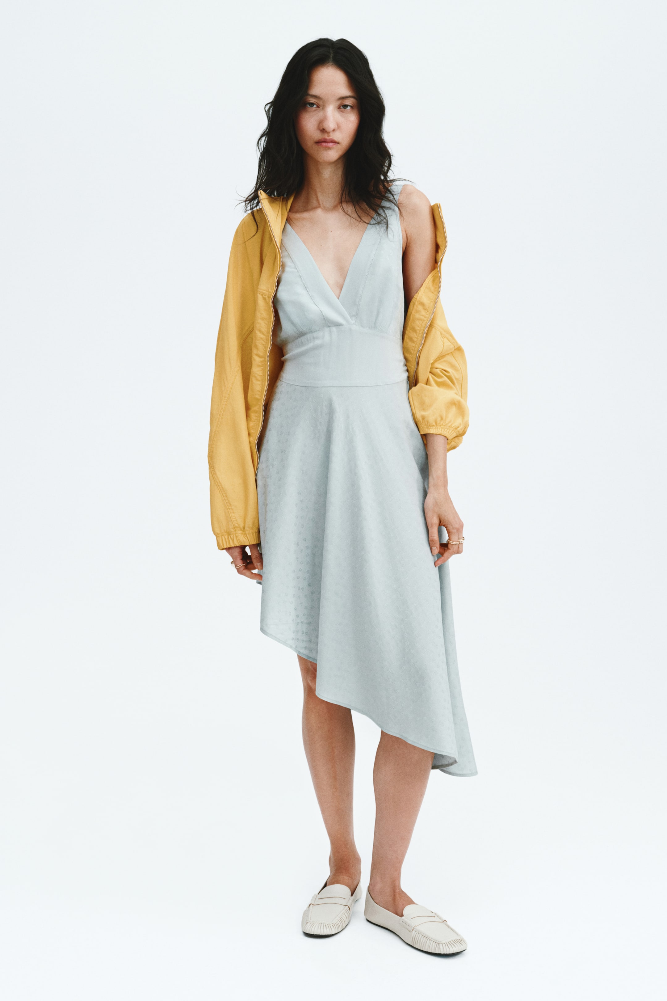 View larger image: A light blue sleeveless dress features a deep V-neckline and a wide fitted waistband, flowing into an asymmetrical skirt with a subtle dotted texture. A golden yellow jacket is draped over the shoulders, and cream loafers complete the look.