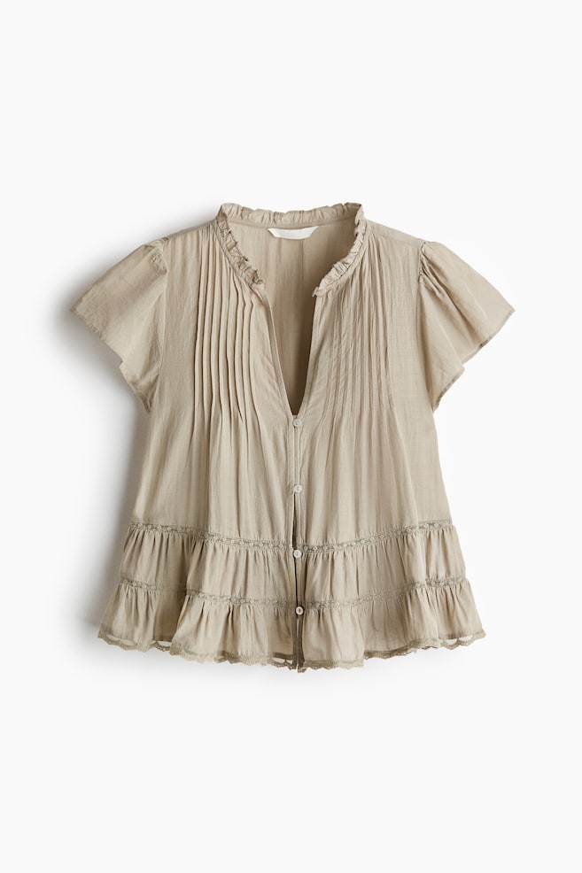 Une blouse beige clair à l'avant présente un col montant à volants et une patte de boutonnage en V partielle. Elle arbore des plis nervurés verticaux sur le corsage, des manches courtes à volants, ainsi qu'un ourlet à étages froncé avec une garniture décorative et des détails en dentelle. - 1