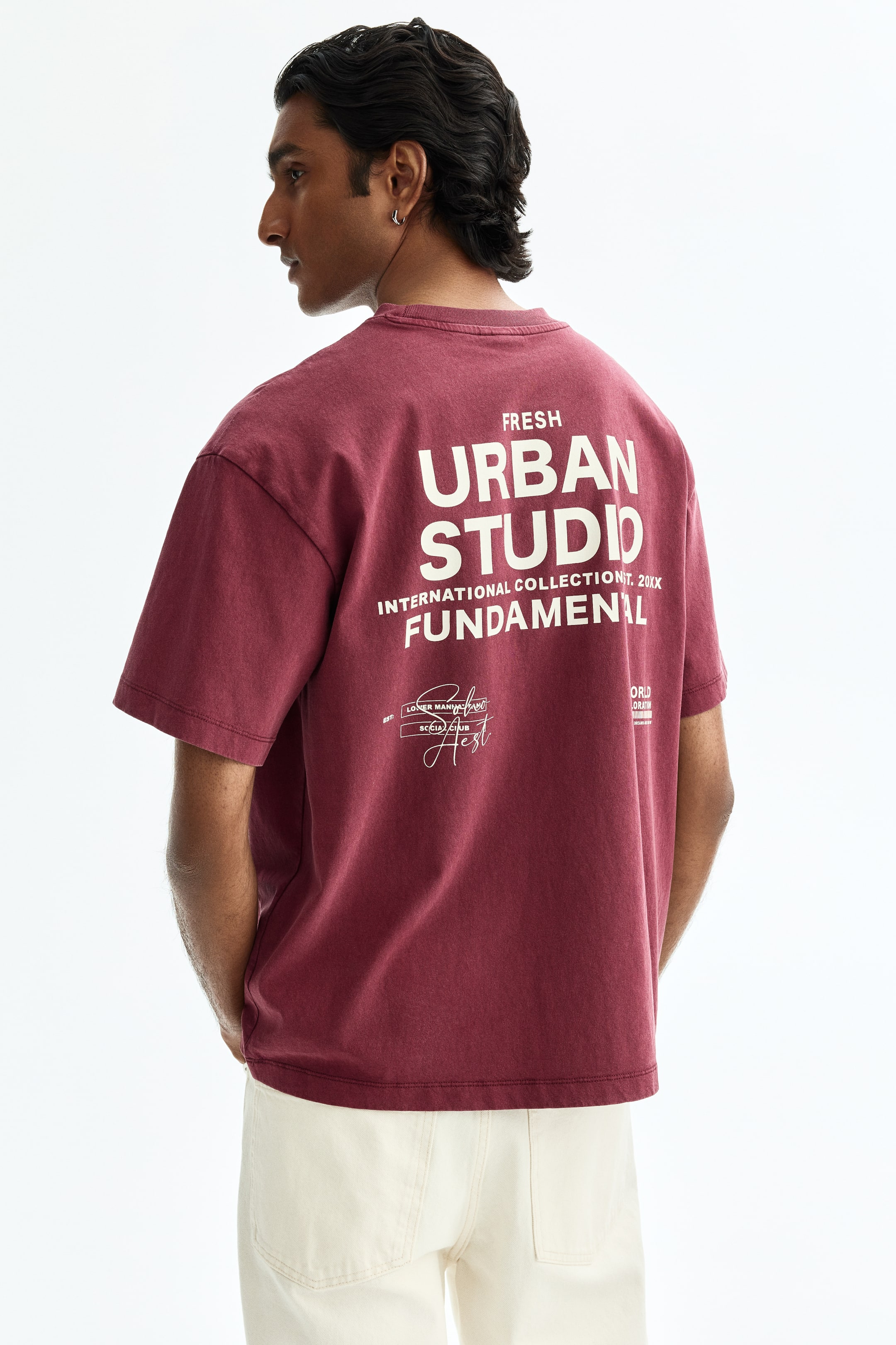 View larger image: A maroon t-shirt is displayed from the back, slightly angled to the right, showcasing a relaxed, oversized fit with short sleeves and a rounded neckline. The back features large white text that reads "FRESH URBAN STUDIO INTERNATIONAL COLLECTION IT. 20XX FUNDAMENTAL", accompanied by smaller white graphic details and text below.