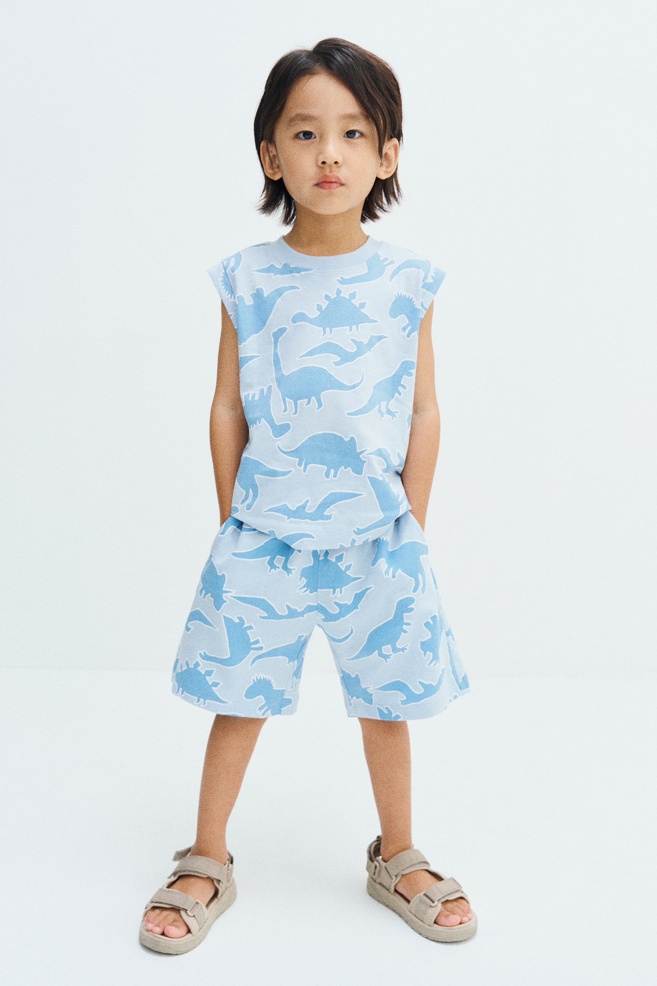 View larger image: The model is wearing knee-length shorts in light blue fabric with an all-over dinosaur print, standing with her hands placed in the side pockets. The shorts are styled as part of a matching set with a sleeveless top in the same print.
