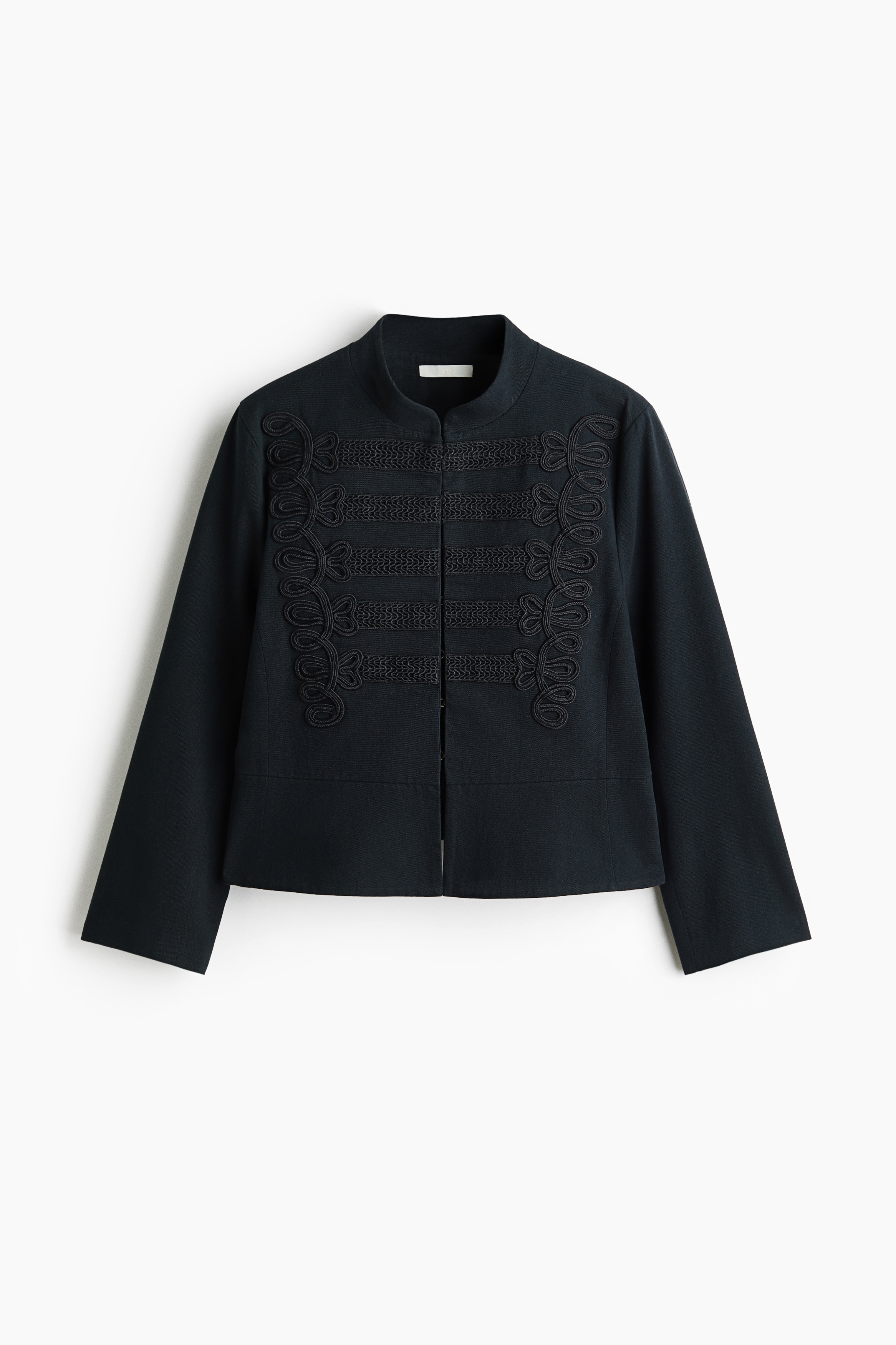 A dark blue, military-style cropped jacket featuring decorative black braided frogging on the front chest and a mandarin collar is being worn by the model. The jacket has three-quarter sleeves and is paired with blue jeans and a black belt.