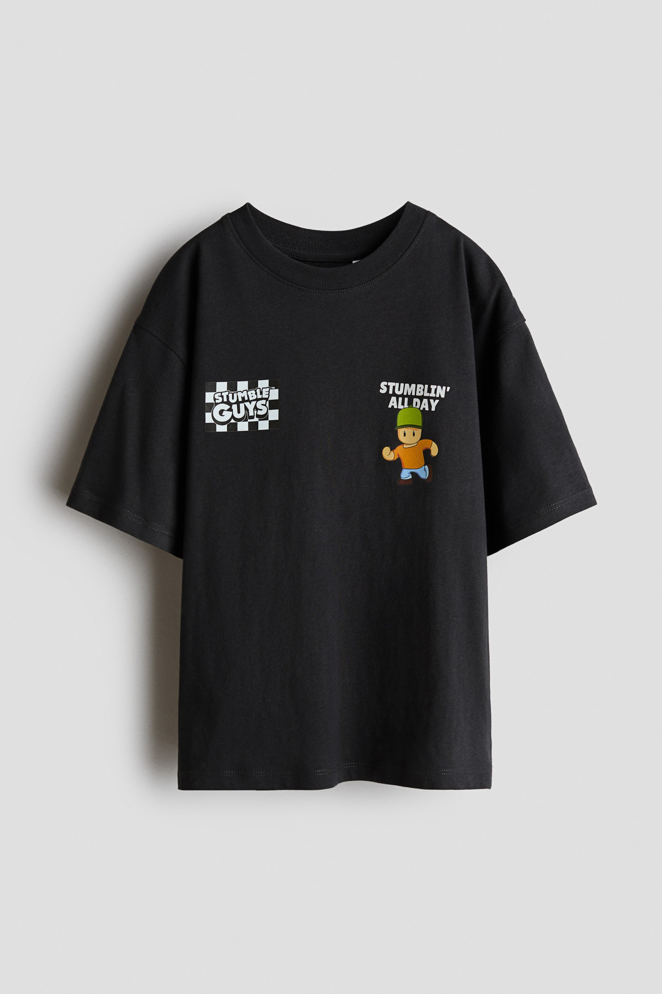 View larger image: A black t-shirt is presenting its front view. It is displaying a white "STUMBLE GUYS" logo over a checkerboard pattern on the left chest, and a cartoon character of a person in a green hat, orange shirt, and blue pants is appearing on the right chest, with "STUMBLIN' ALL DAY" text visible above it.