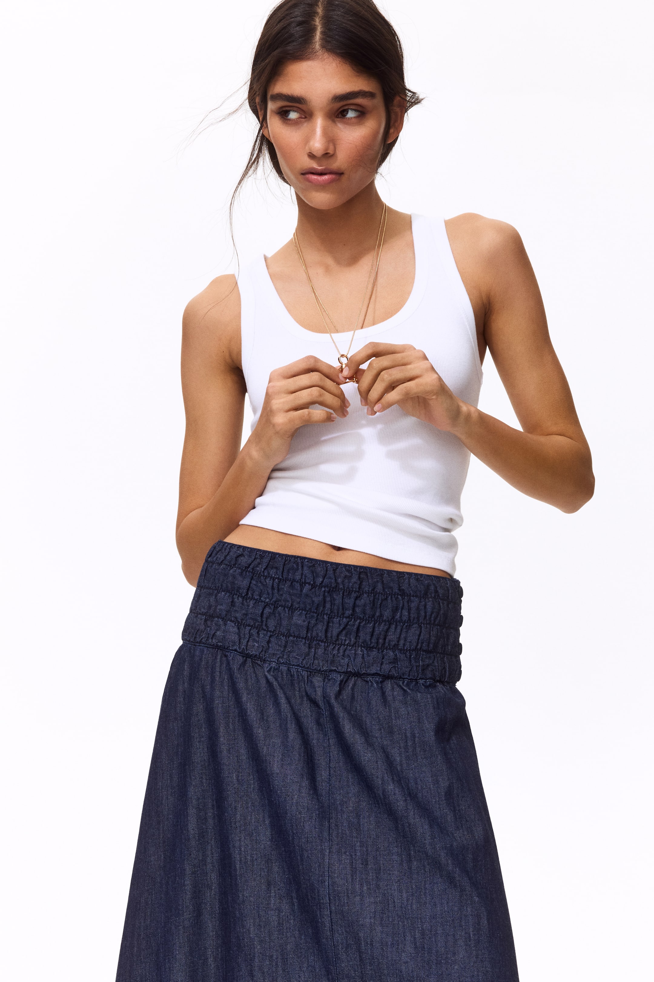 View larger image: The model dons a calf-length, circular skirt crafted from dark wash cotton denim, showcasing a high waist with broad, smocked elastication. Complementing the skirt, the model is styled in a white ribbed tank top.